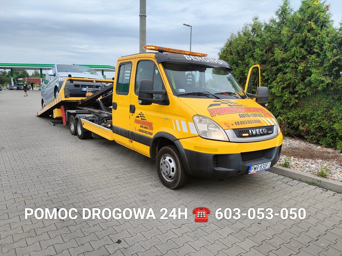 Żółta laweta Iveco z załadowanym busem na parkingu. Widoczny numer telefonu alarmowego i logo firmy. Pomoc drogowa 24h, szybka interwencja.