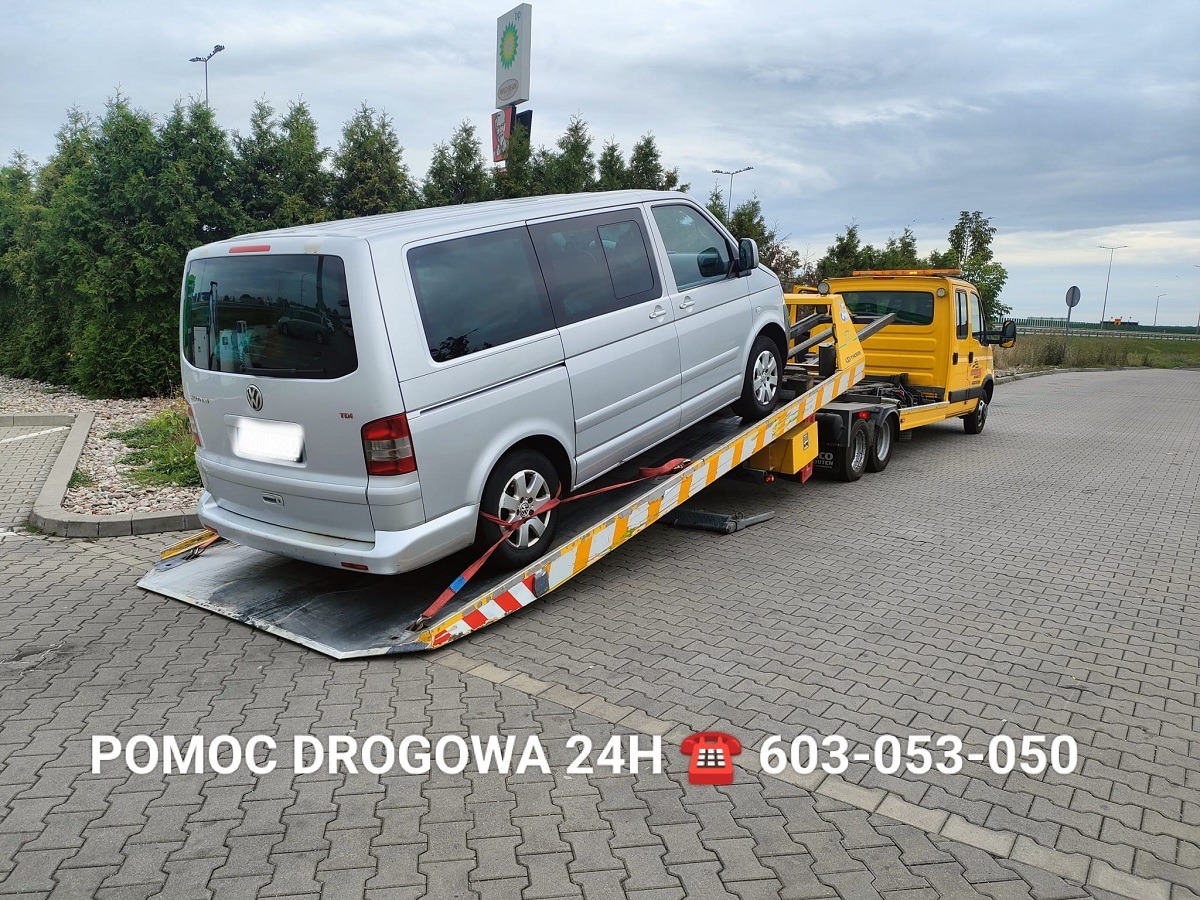 Żółta laweta z srebrnym busem VW na platformie, zabezpieczonym pasami. Widok z tyłu, w tle drzewa i znak BP. Na dole napis 'Pomoc Drogowa 24H' i numer telefonu.