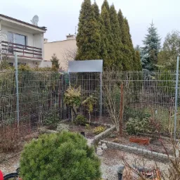 Panelowa siatka ogrodzeniowa z widocznymi słupkami na tle zadbanego ogrodu z tujami i niskimi krzewami. W tle widoczny budynek mieszkalny.