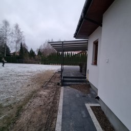 Karo-Met - Nowoczesna pergola z metalową konstrukcją przylegająca do białego domu z widocznymi schodami i ścieżką z kostki brukowej, w tle trawnik pokryty resztkami śniegu i zimowe drzewa.