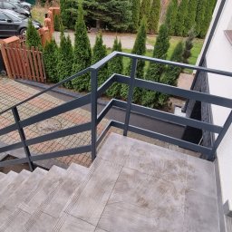 Karo-Met - Zewnętrzne schody z szarymi płytkami i nowoczesną metalową balustradą w kolorze grafitowym, widok z góry na tle zielonych tuj i kostki brukowej.