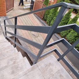 Karo-Met - Zewnętrzne schody wyłożone płytkami z szarą metalową balustradą, widok z góry na dół, z fragmentem chodnika z kostki i tujami w tle.