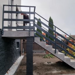 Karo-Met - Zewnętrzne schody z metalową balustradą prowadzące do podwyższonego wejścia, betonowe stopnie, szara kolorystyka, widok z boku na tle domu i ogrodu.