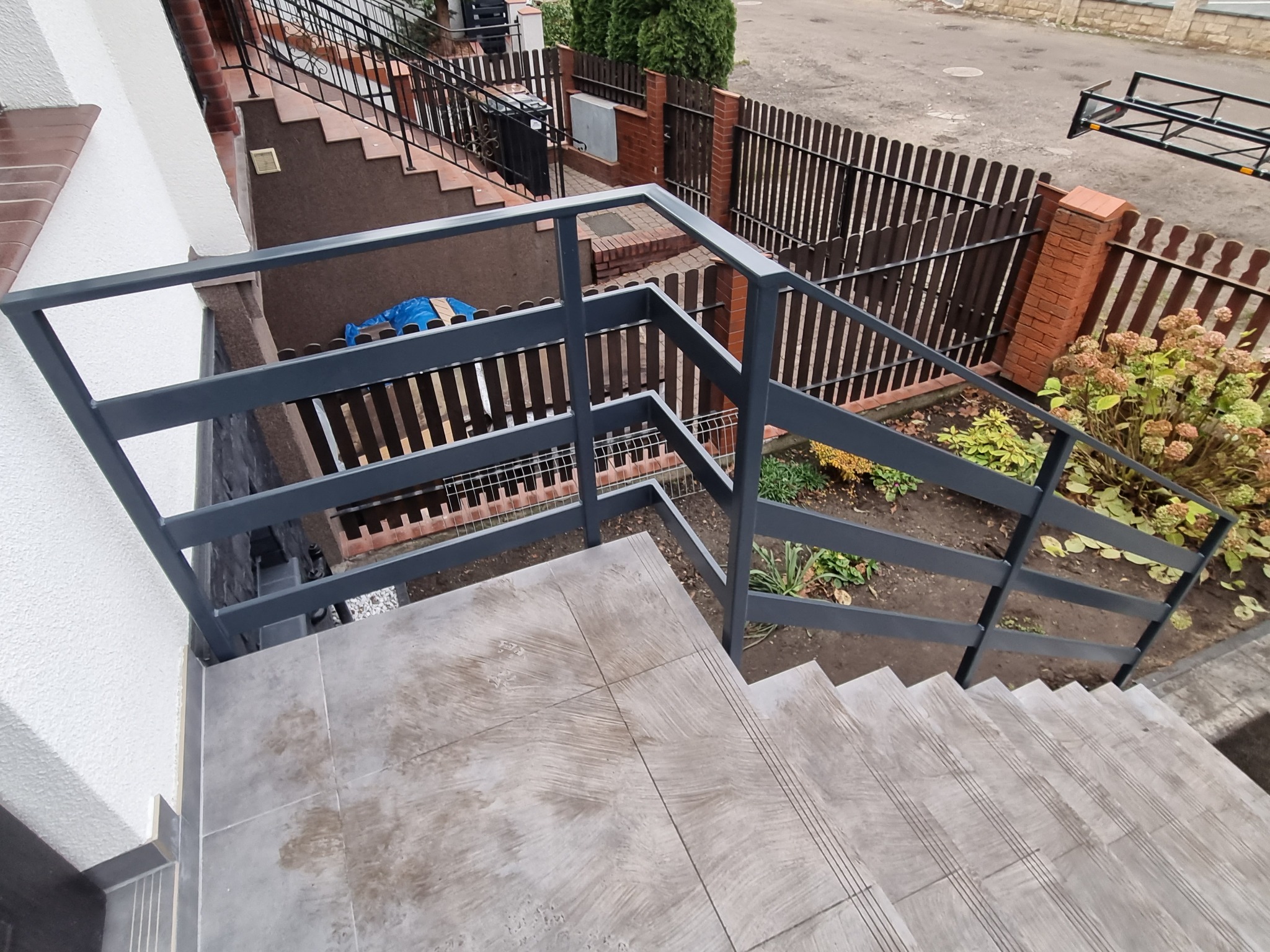 Zewnętrzne schody z szarymi, metalowymi barierkami, wyłożone płytkami imitującymi drewno, prowadzące do wejścia do budynku.