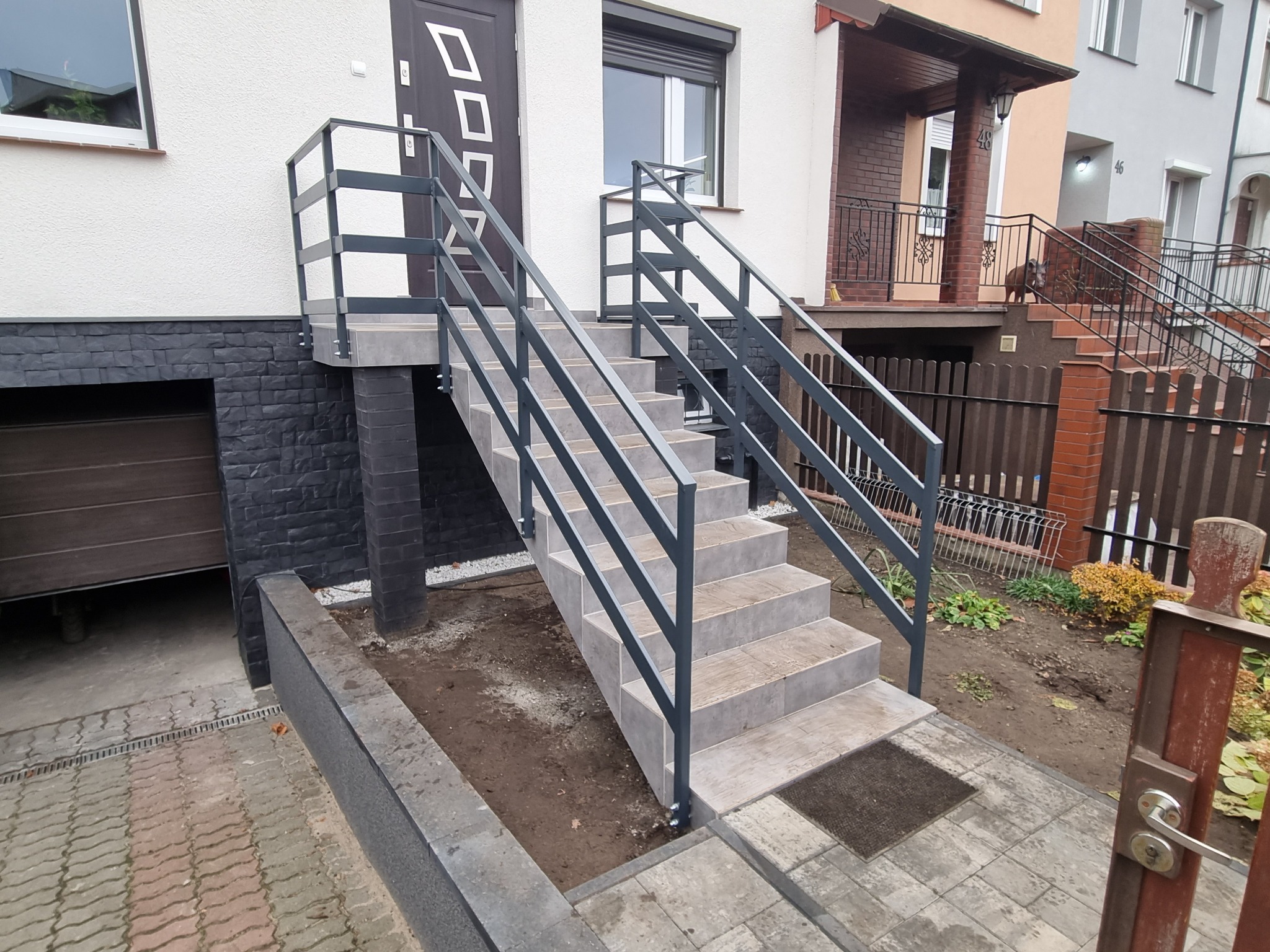 Zewnętrzne schody z metalową balustradą prowadzące do drzwi wejściowych, wykończone płytkami ceramicznymi, z widocznym fragmentem garażu po lewej stronie.
