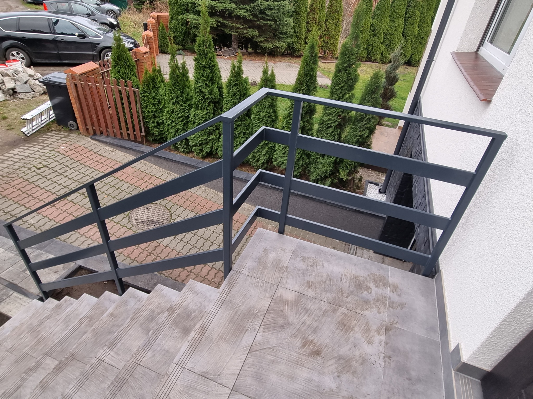 Zewnętrzne schody z szarymi płytkami i nowoczesną metalową balustradą w kolorze grafitowym, widok z góry na tle zielonych tuj i kostki brukowej.