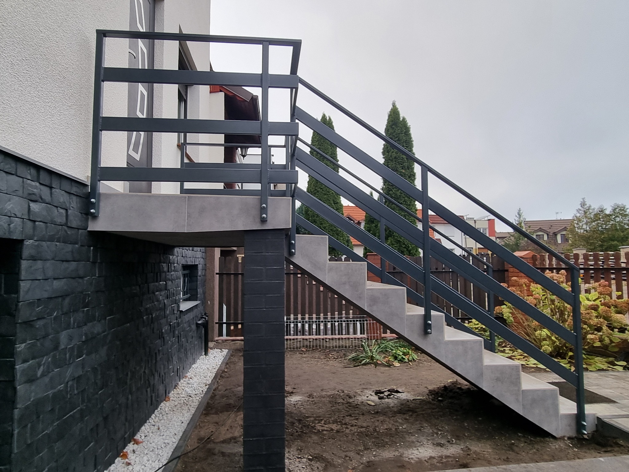 Zewnętrzne schody z metalową balustradą prowadzące do podwyższonego wejścia, betonowe stopnie, szara kolorystyka, widok z boku na tle domu i ogrodu.