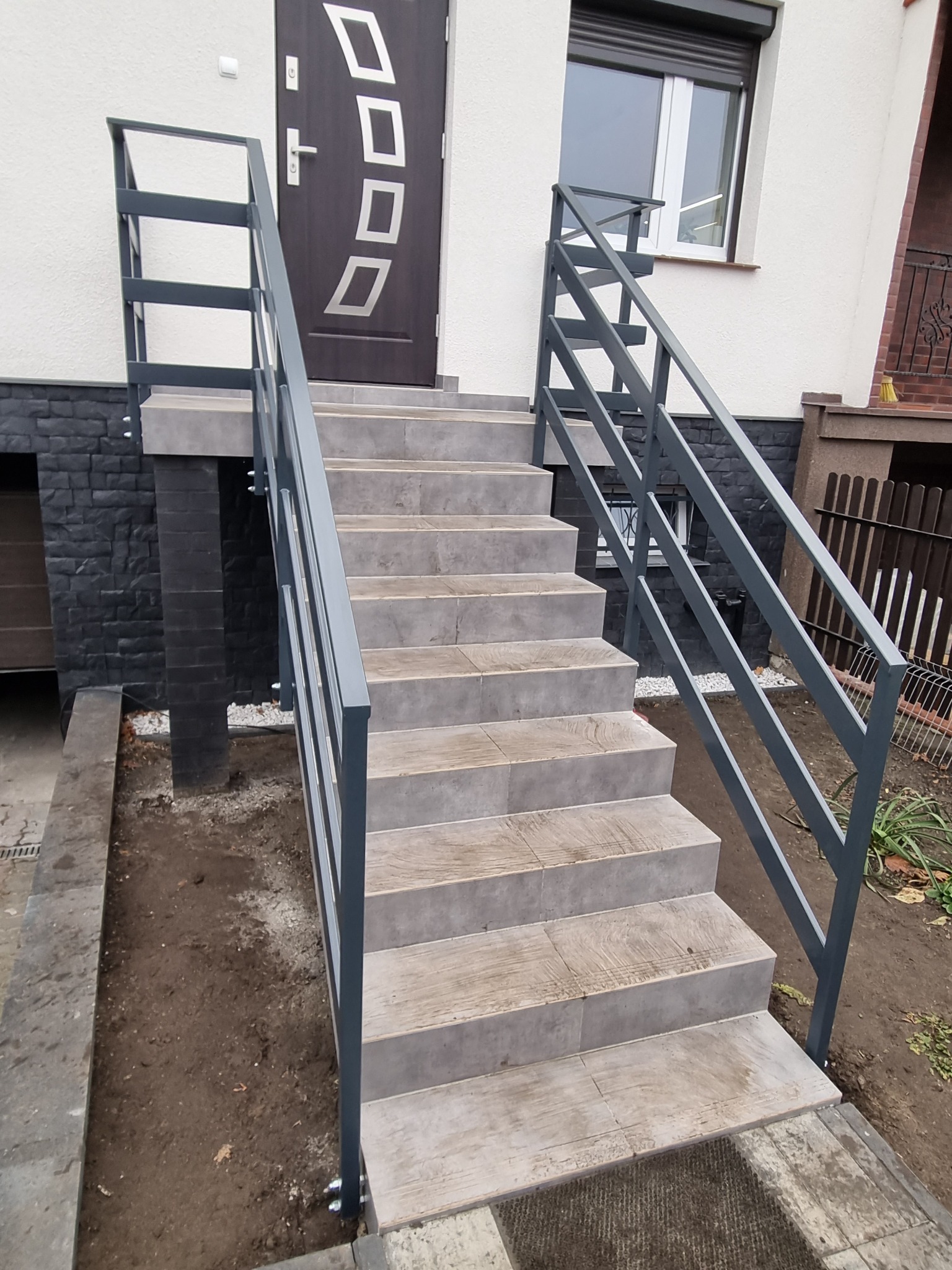 Zewnętrzne schody z szarego metalu i płytek prowadzące do drzwi wejściowych z geometrycznym wzorem, widoczne również okno z roletą.