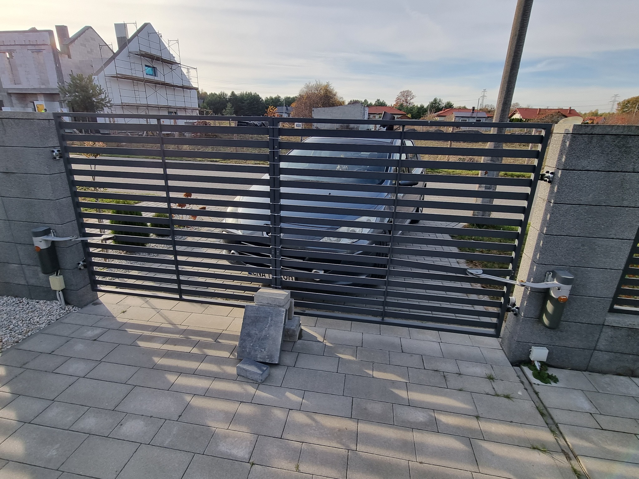 Nowoczesna, pozioma brama wjazdowa z automatyką, widok przez szare, poziome profile na zaparkowany samochód, kostka brukowa i budynek w budowie w tle.