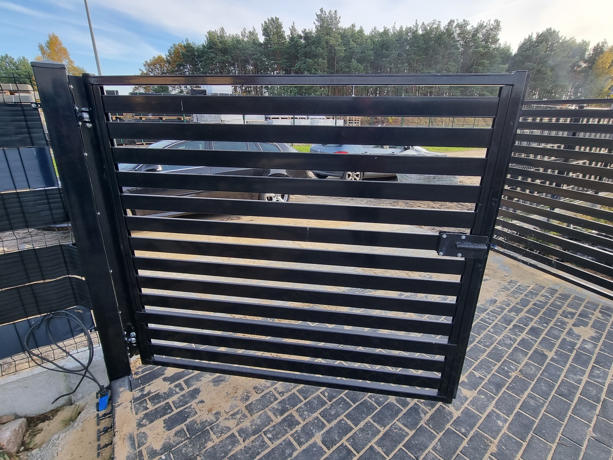 Czarna, pozioma brama panelowa otwarta częściowo, widoczna kostka brukowa na podjeździe, w tle zaparkowane samochody i drzewa.