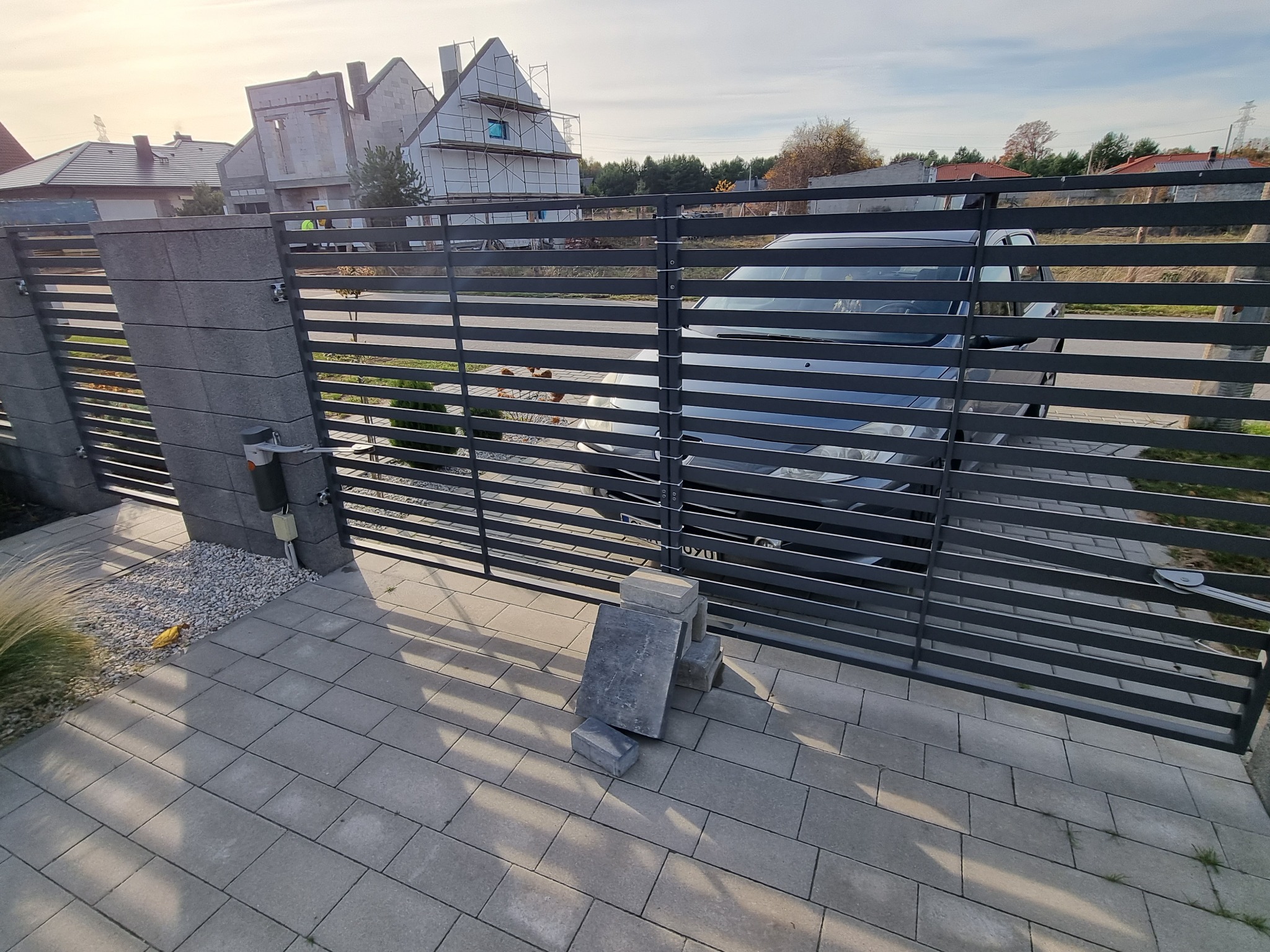 Nowoczesna, pozioma brama wjazdowa z automatyką, widok z zewnątrz, na posesję wjeżdża samochód, obok kostka brukowa i elementy montażowe.