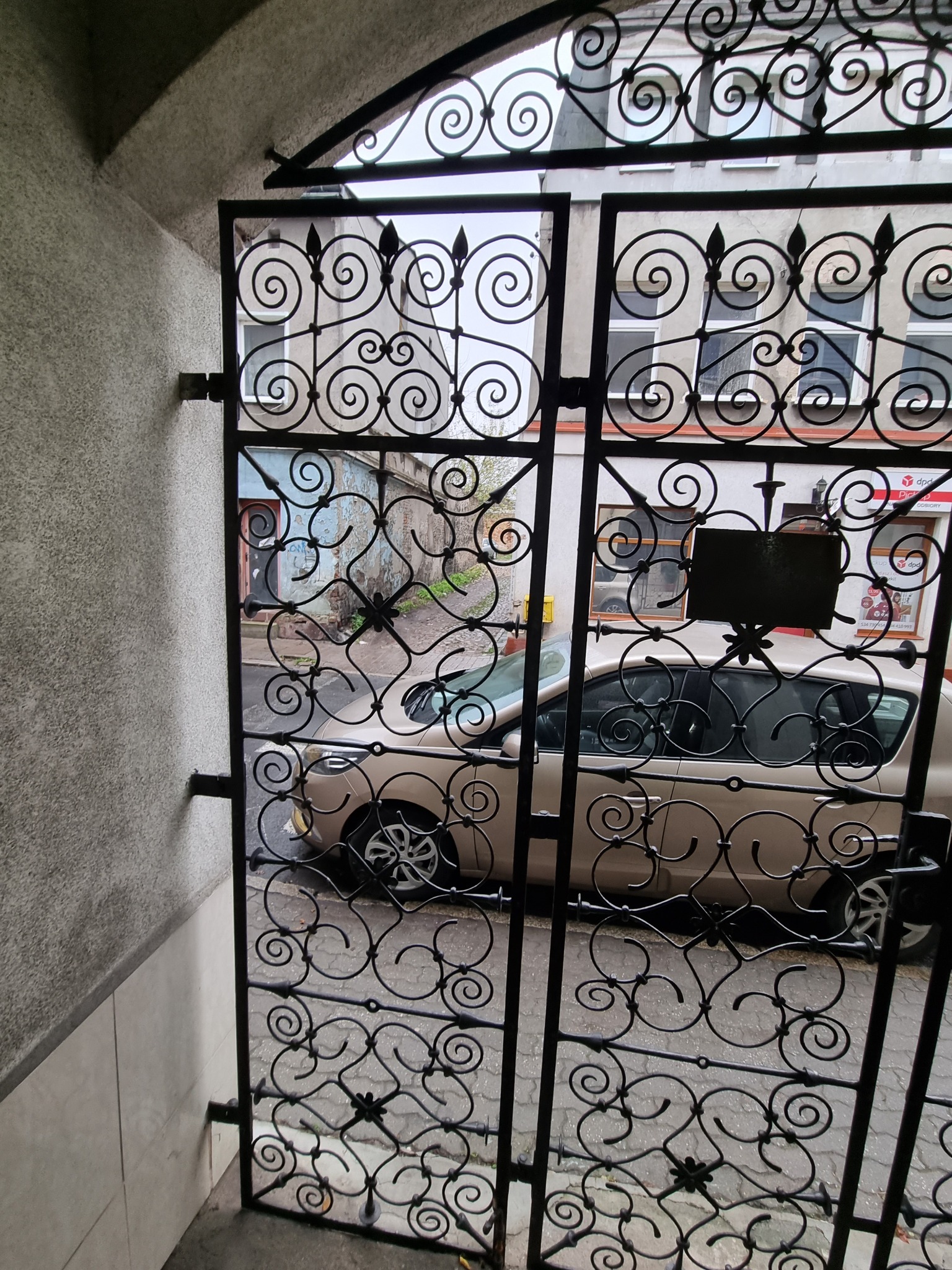 Otwarta, ozdobna metalowa brama z motywem spiralnym, widok na ulicę z zaparkowanym samochodem i budynki w tle.