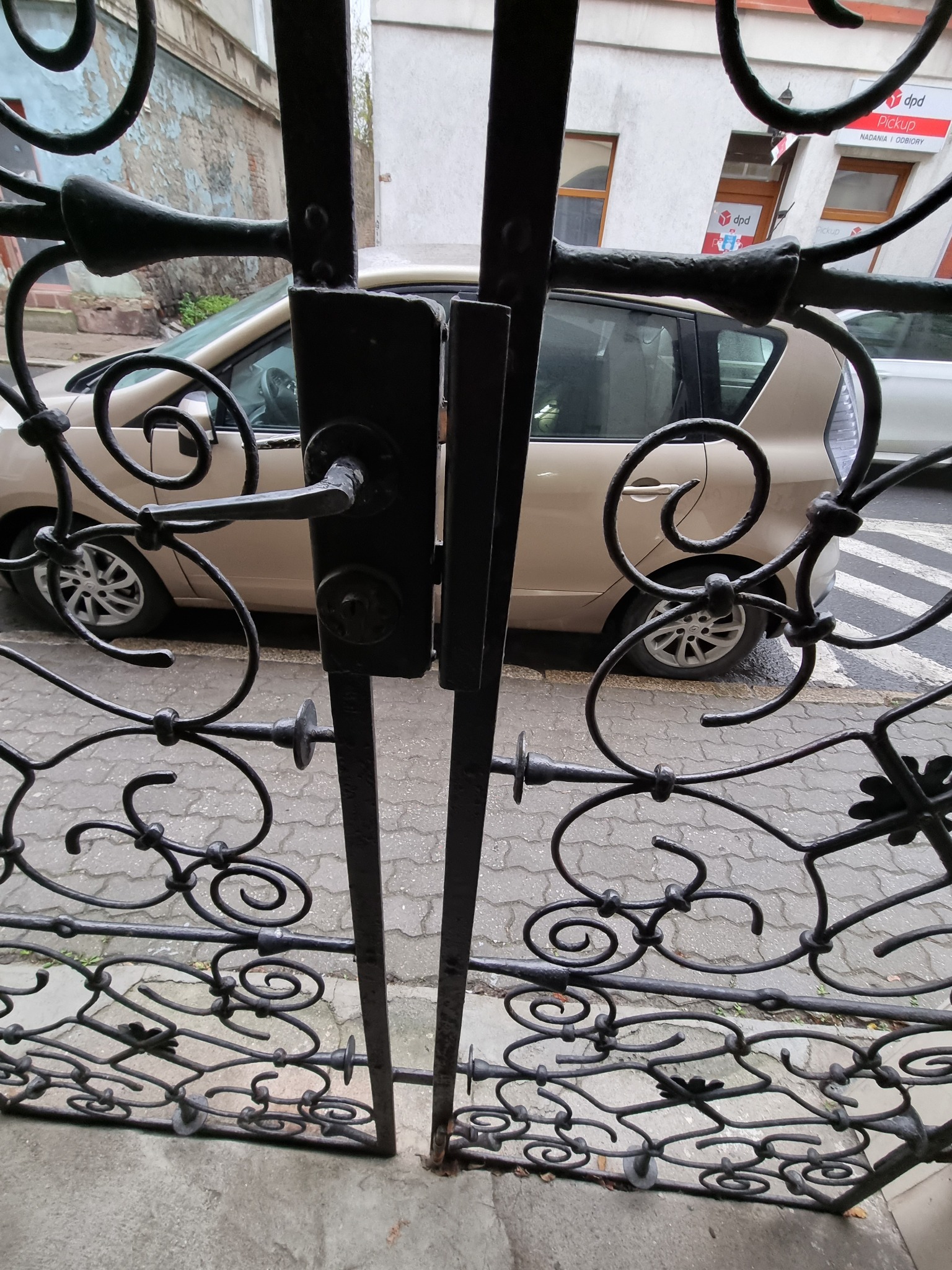 Kute, metalowe ogrodzenie bramy z ozdobnymi elementami, widok z bliska na zamek i klamkę, w tle zaparkowany samochód i budynek z szyldem kurierskim.