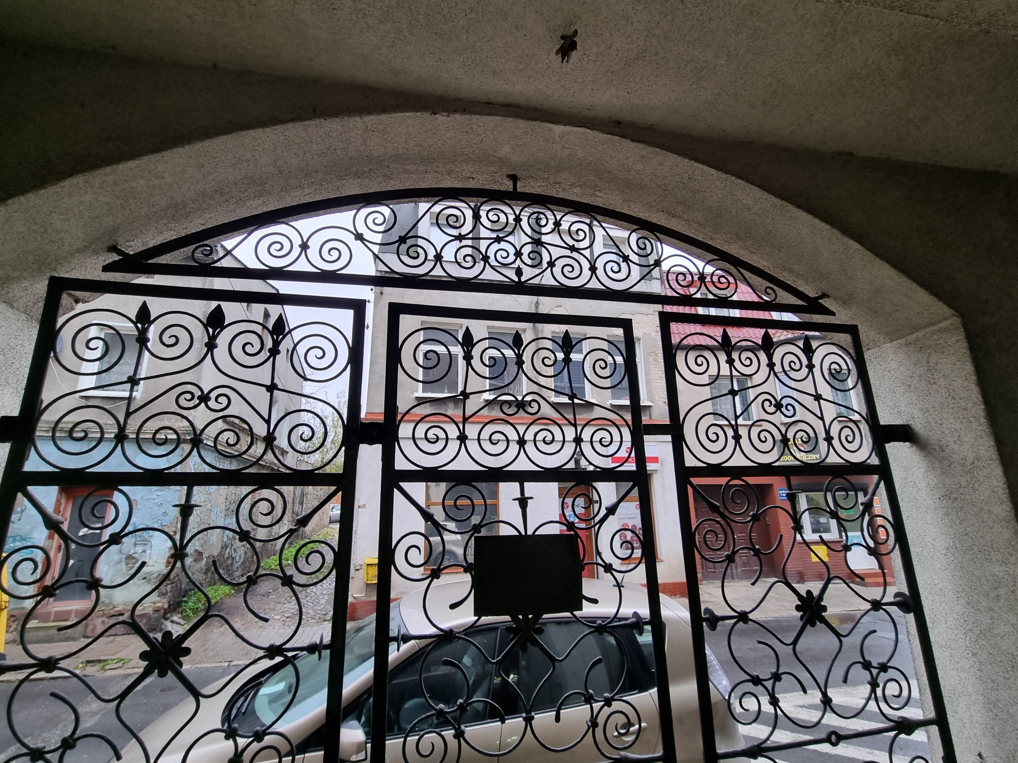 Otwarta, ozdobna brama metalowa z kręconymi elementami, widziana z perspektywy wnętrza przejścia, z widokiem na ulicę i zaparkowany samochód.