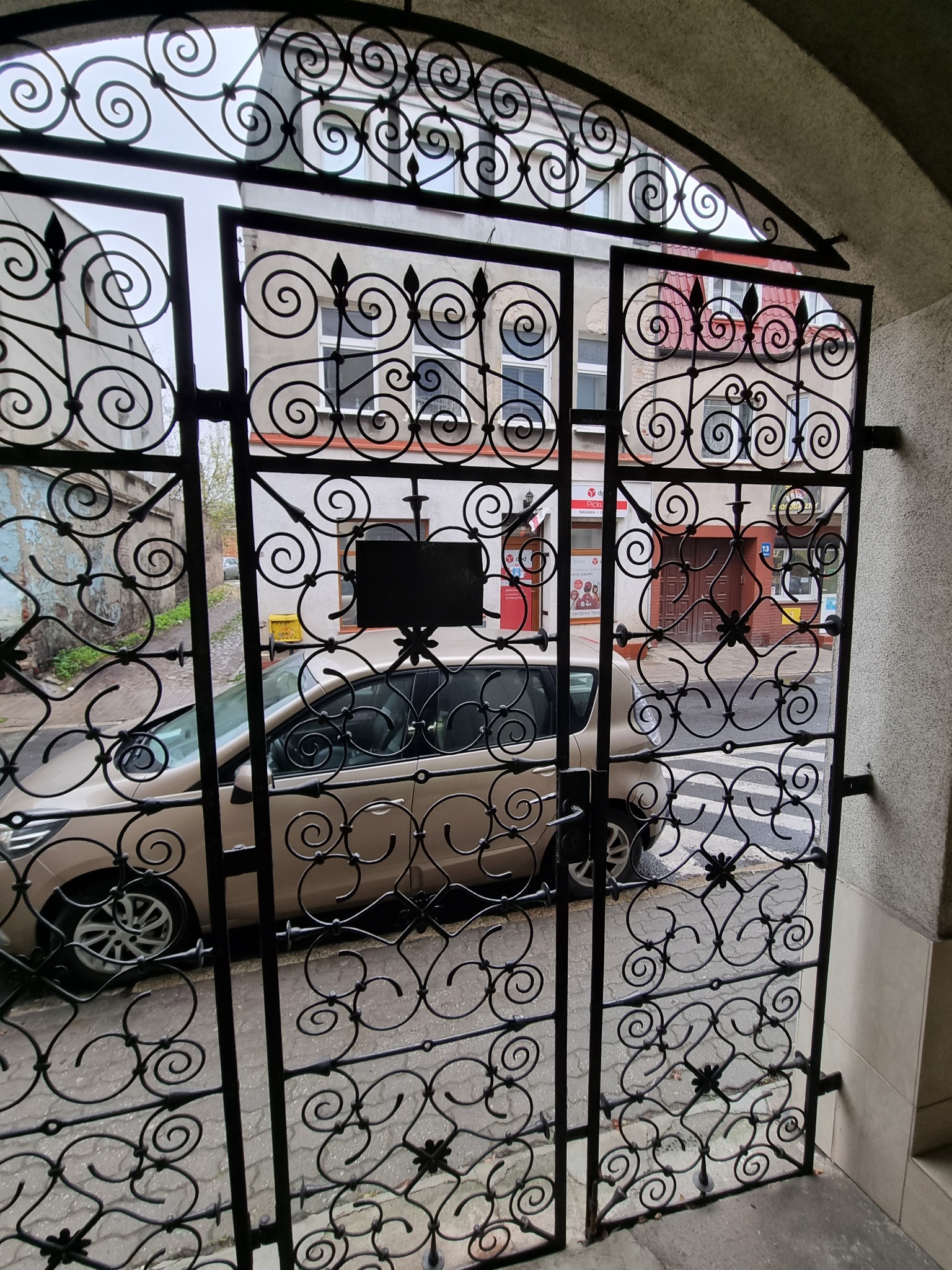 Ozdobna, metalowa brama wejściowa z kutymi elementami w kształcie spiral, widziana od wewnątrz budynku, z widokiem na ulicę i zaparkowany samochód.