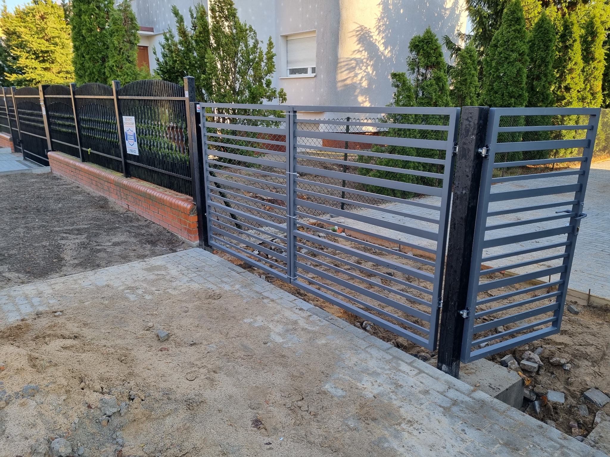 Nowoczesna, pozioma brama w kolorze szarym, z widocznym mechanizmem zamykania, zamontowana między dwoma czarnymi słupkami, z fragmentem ogrodzenia panelowego w tle, na tle trawnika i budynku...