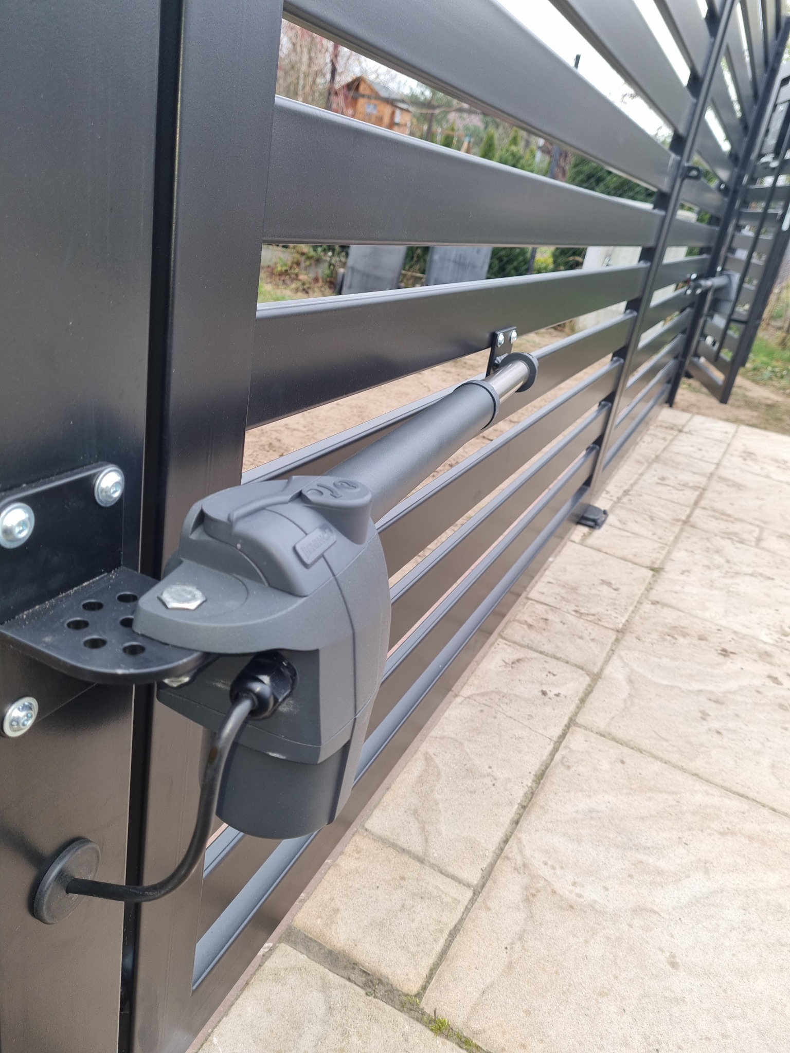 Szczegółowe ujęcie automatycznego napędu do bramy przesuwnej, zamontowanego na nowoczesnym, poziomym ogrodzeniu z metalowych profili w kolorze antracytowym.