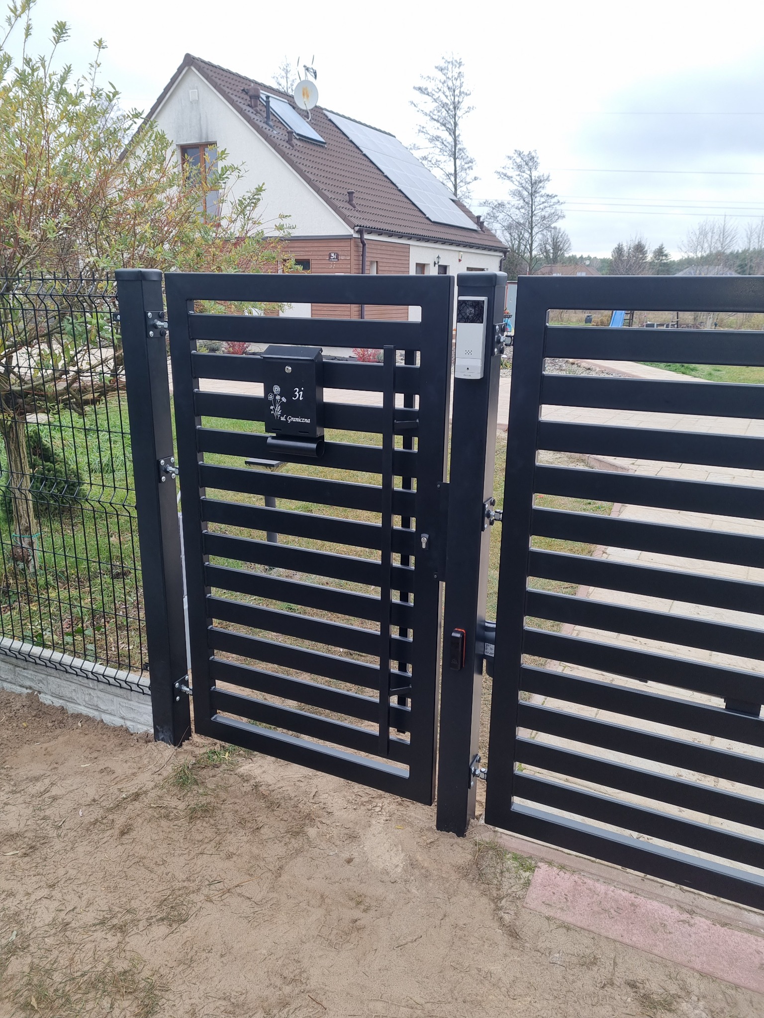 Nowoczesna, czarna brama posesji z poziomymi szczeblami, skrzynką na listy z adresem i domofonem, na tle domu z panelami słonecznymi.