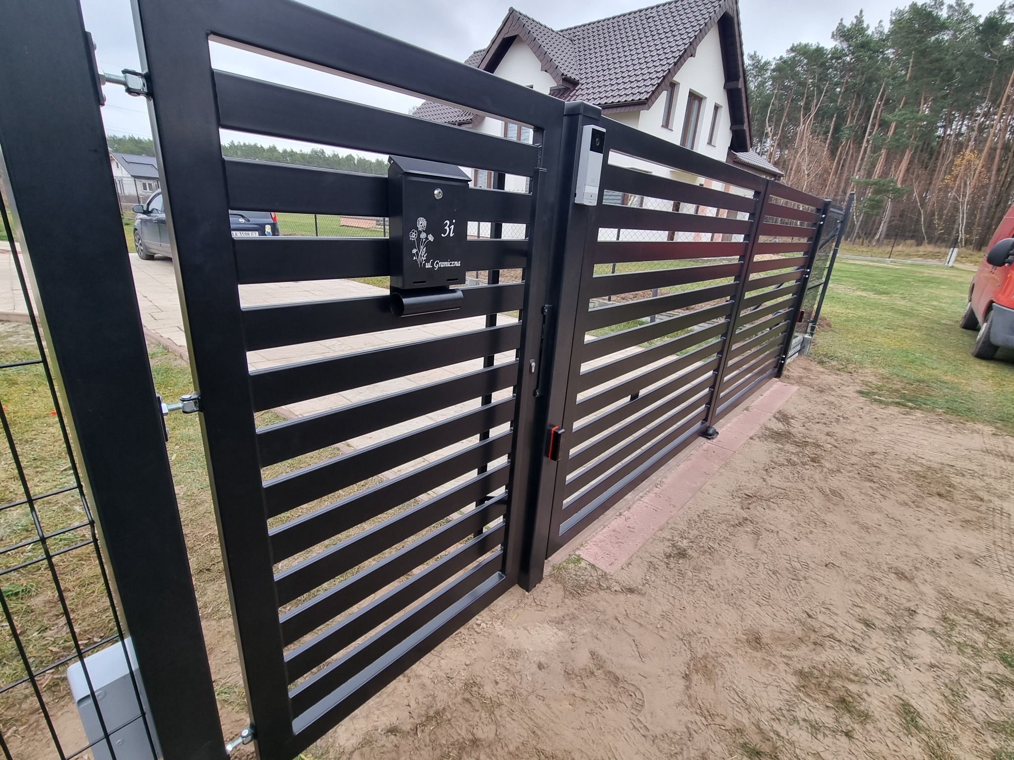 Nowoczesna, pozioma brama wjazdowa z czarnego metalu z zamontowaną skrzynką na listy i domofonem, Wolwark. W tle dom z brązowym dachem i drzewa.