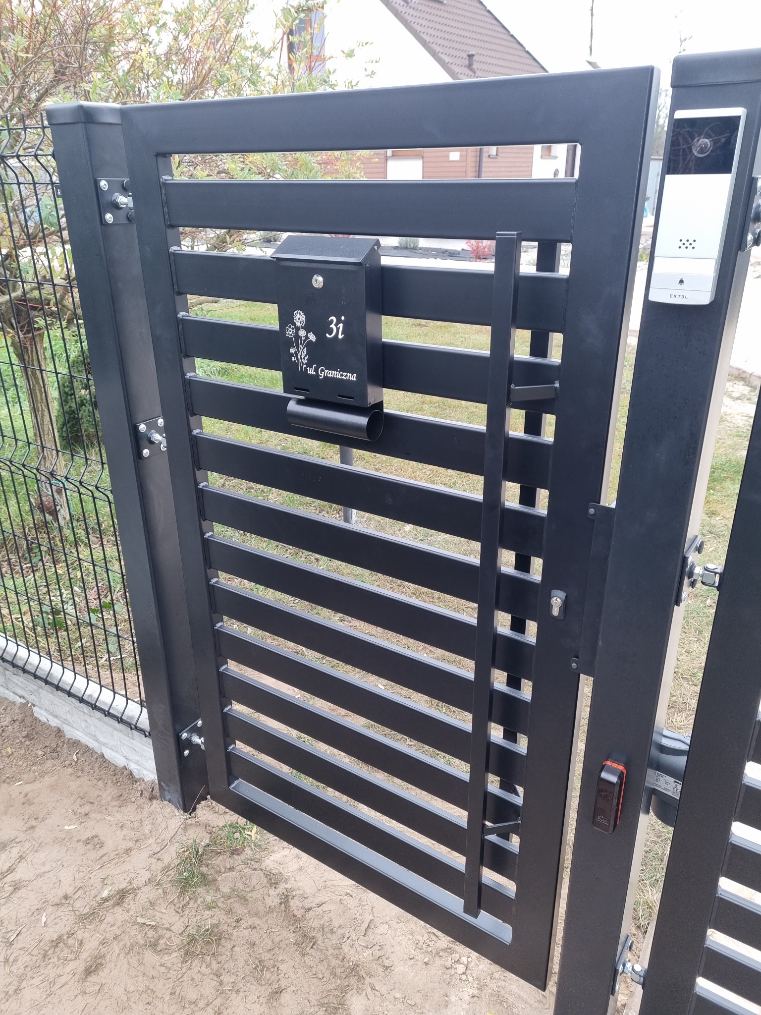 Nowoczesna, czarna brama posesyjna z poziomymi szczeblami, wyposażona w skrzynkę na listy z adresem 'ul. Graniczna 3i', domofon i automat otwierający, widoczna na tle siatki ogrodzeniowej.