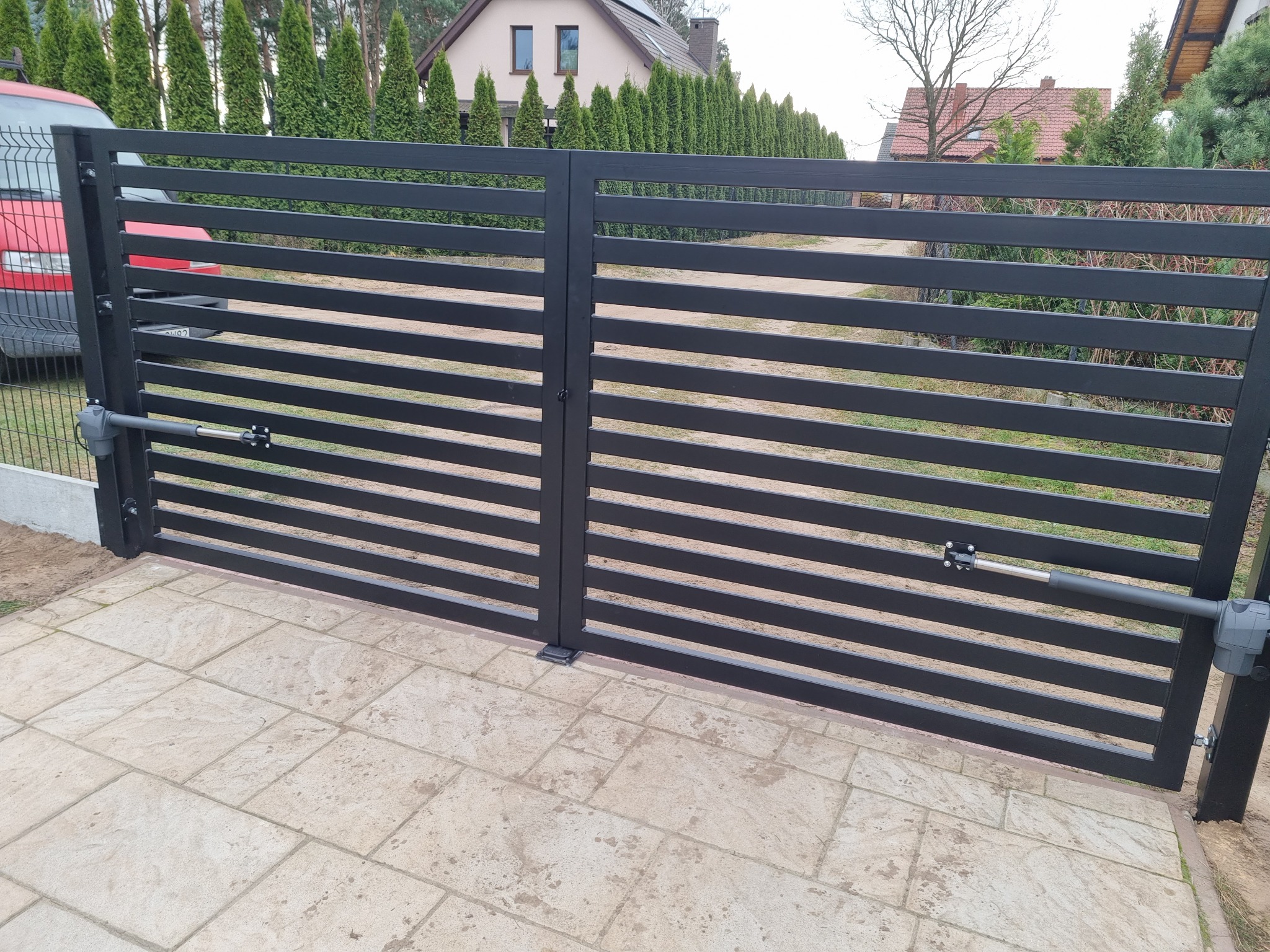 Nowoczesna, pozioma brama dwuskrzydłowa w kolorze antracytowym z automatyką, zamontowana na posesji z kostką brukową i tujami w tle.