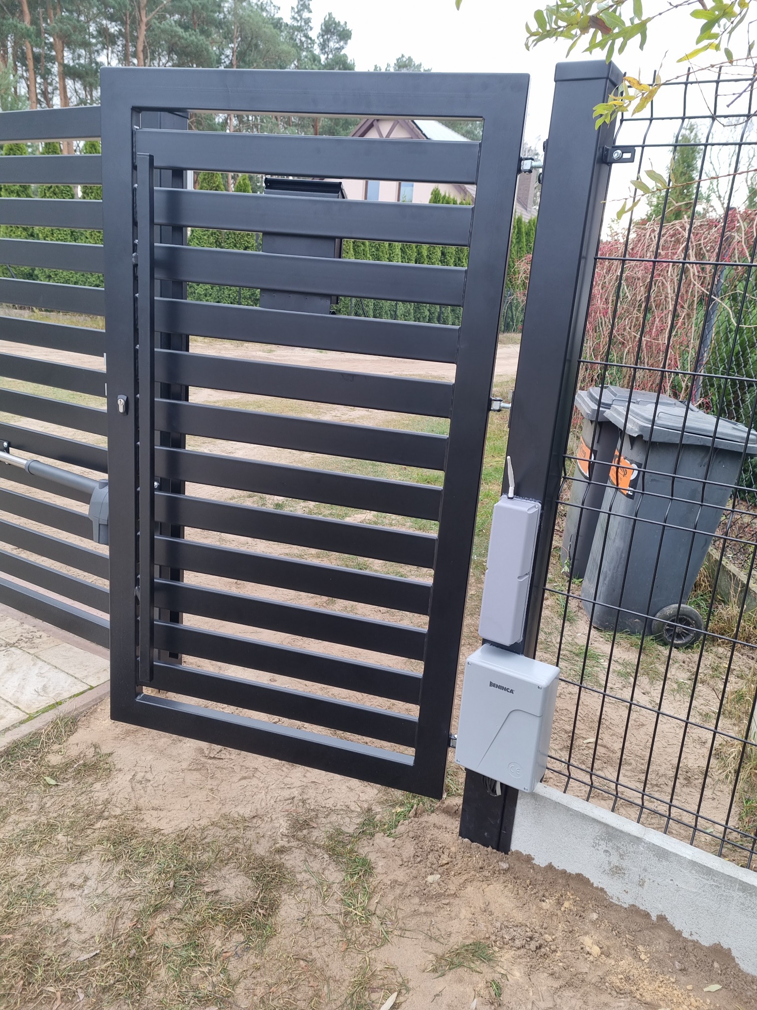 Nowoczesna, pozioma, metalowa brama w kolorze antracytowym z automatyką Beninca, zamontowana przy ogrodzeniu z siatki, na tle domu i zieleni.