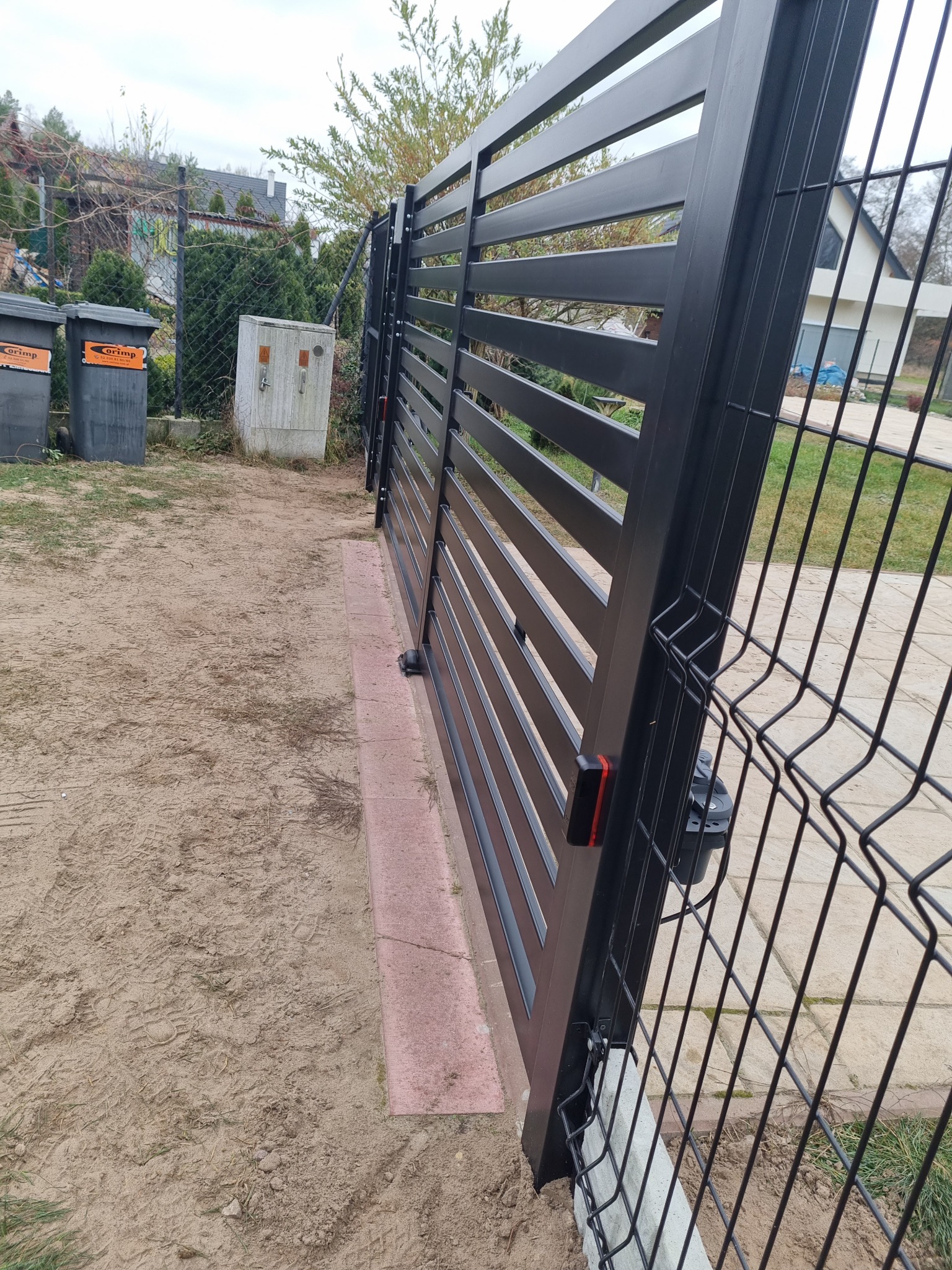 Nowoczesna brama przesuwna z poziomych profili stalowych w kolorze grafitowym, widoczny napęd automatyczny i fotokomórka, z boku fragment ogrodzenia panelowego z siatki.