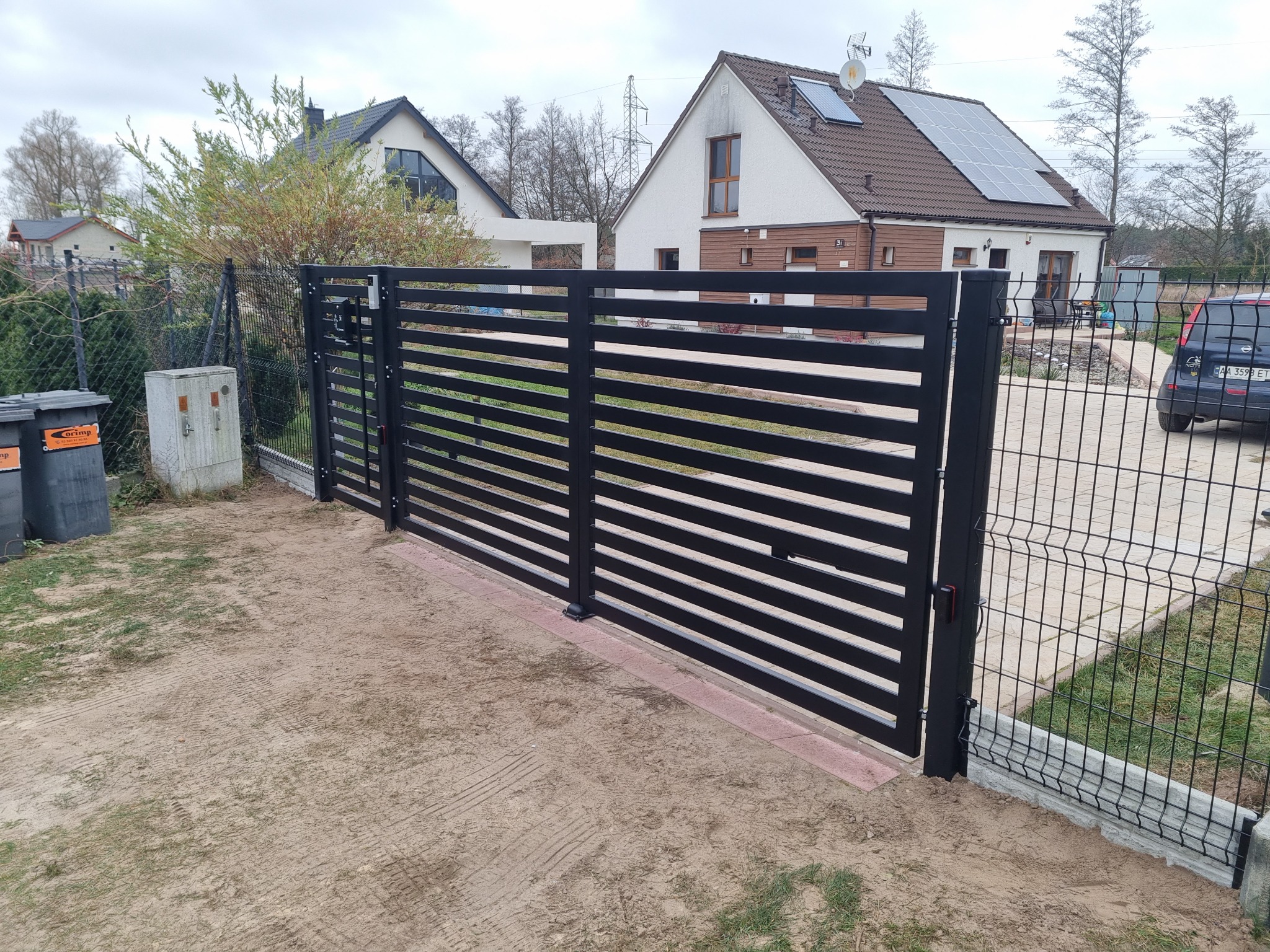 Nowoczesna, pozioma brama wjazdowa w kolorze czarnym, zintegrowana z ogrodzeniem z siatki, na tle domu z panelami słonecznymi.