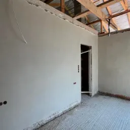 Świeżo położone, gładkie szare tynki na ścianach w surowym wnętrzu domu w trakcie budowy, widoczne otwory na gniazdka elektryczne i surowy strop z drewnianych belek.