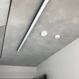 Długa, prostokątna lampa LED z czarną obudową zamontowana na szarym, surowym betonowym suficie, obok dwa okrągłe czujniki dymu lub ruchu, widok z dołu.