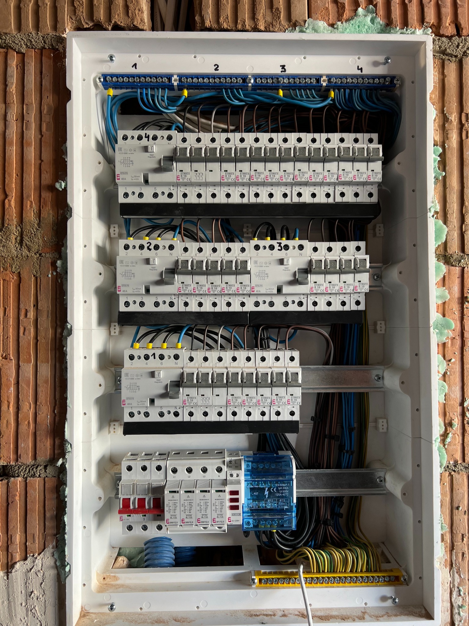 Rozdzielnica elektryczna w trakcie montażu, widoczne zabezpieczenia różnicowoprądowe, bezpieczniki nadprądowe, listwy zaciskowe i przewody w kolorach niebieskim, czarnym i żółto-zielonym...