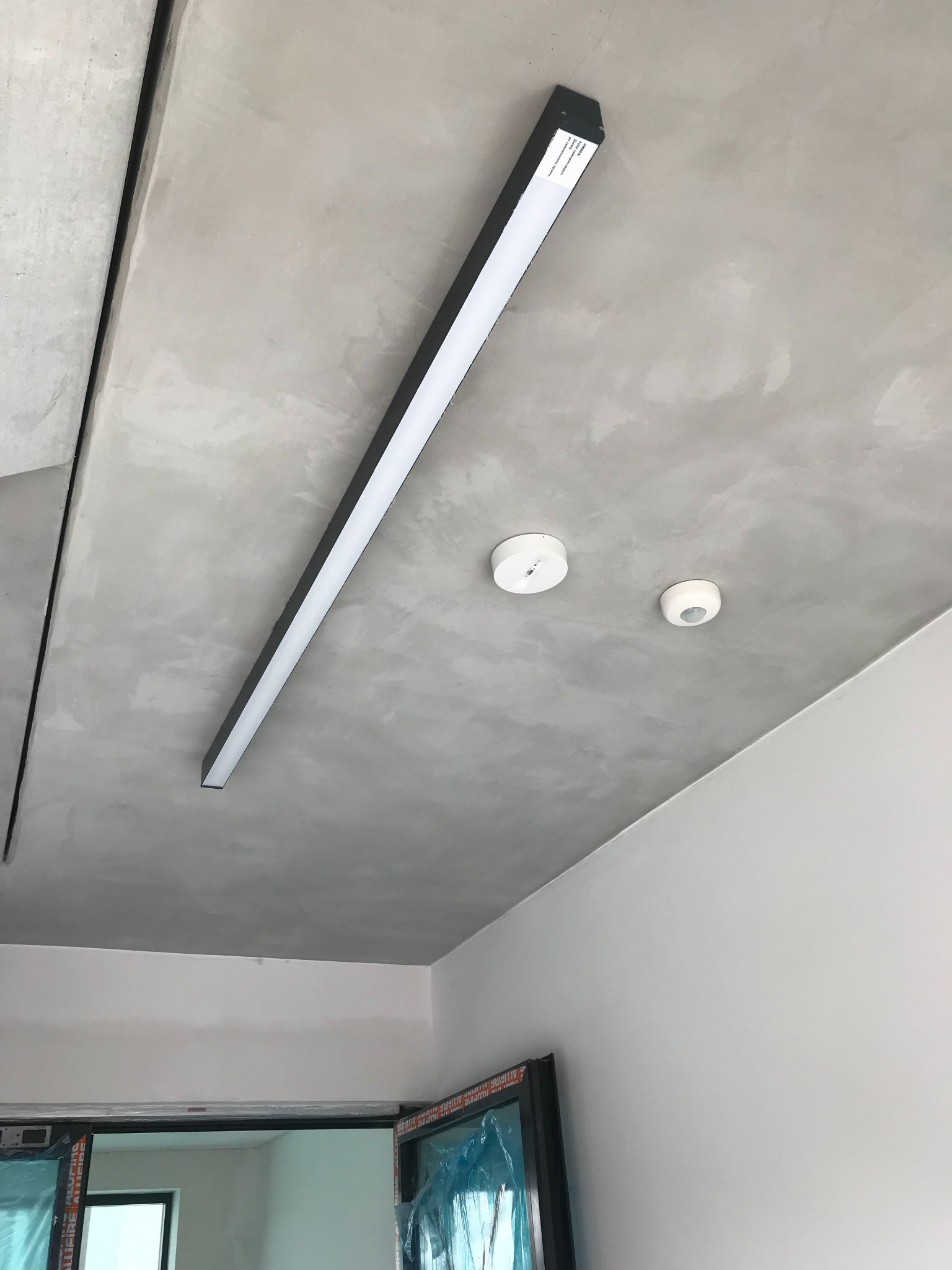 Długa, prostokątna lampa LED z czarną obudową zamontowana na szarym, surowym betonowym suficie, obok dwa okrągłe czujniki dymu lub ruchu, widok z dołu.