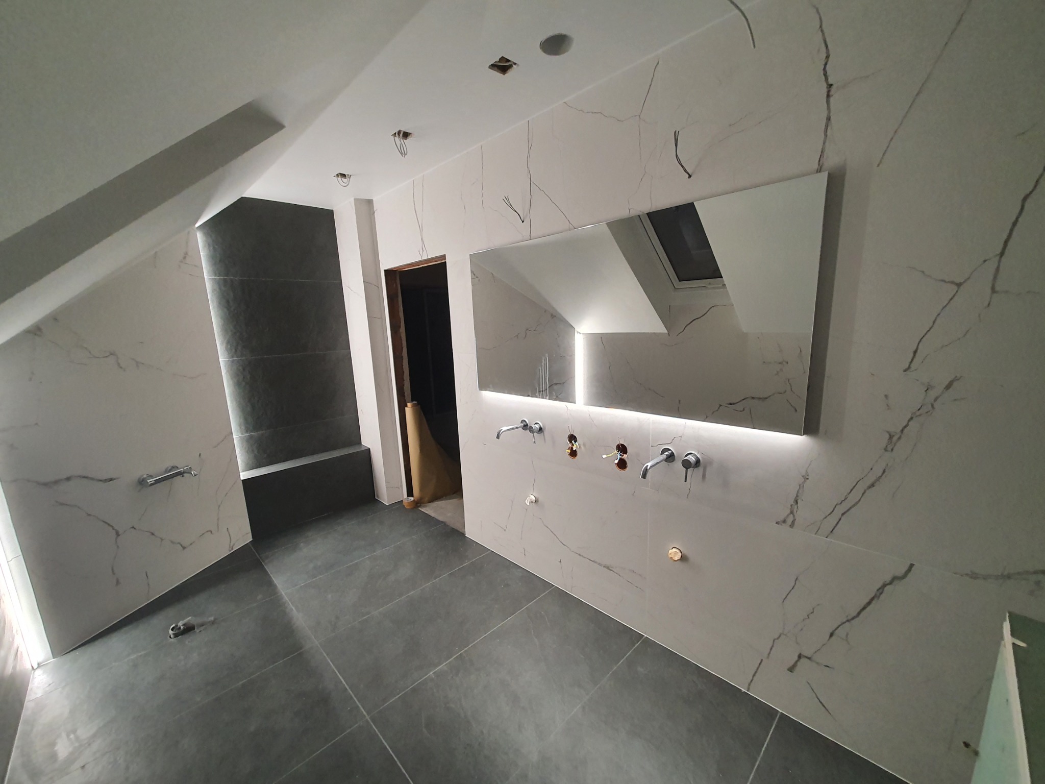 Nowoczesna łazienka na poddaszu w trakcie wykańczania, szare płytki podłogowe, ściany wyłożone białymi płytkami imitującymi marmur, widoczne punkty instalacji wodnej i elektrycznej, lustro...