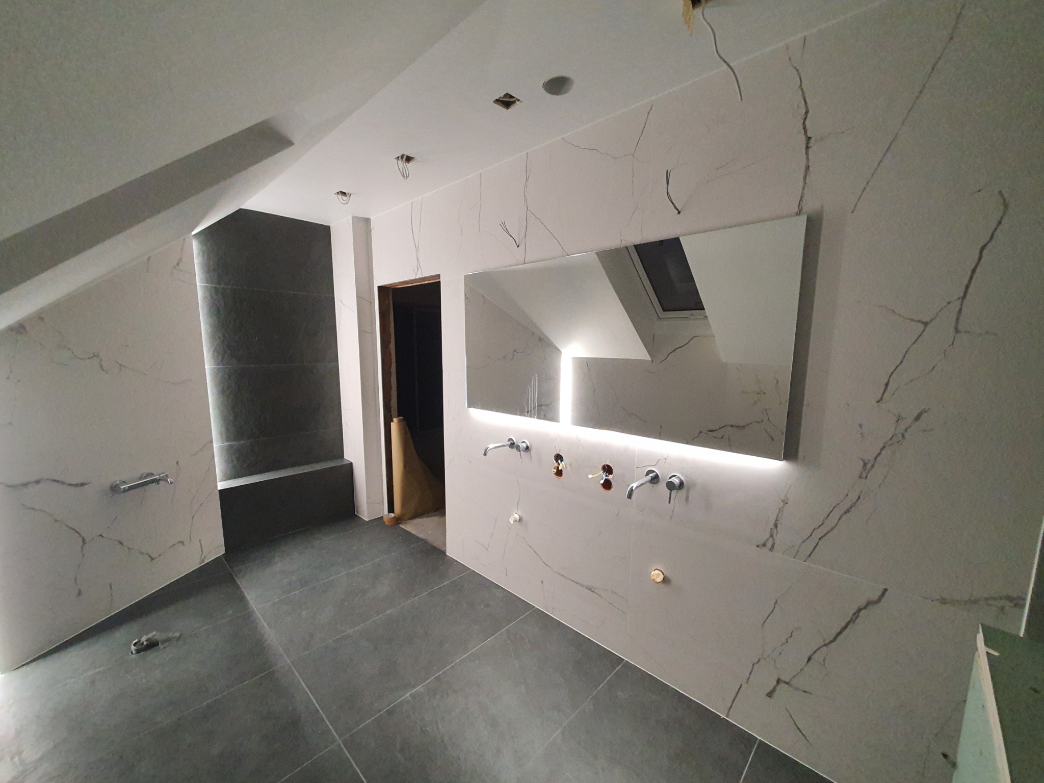Łazienka w trakcie wykańczania w poddaszu: szare płytki podłogowe, ściany wyłożone płytkami imitującymi marmur z widocznymi otworami na baterie umywalkowe, lustro z podświetleniem LED, widoczne...