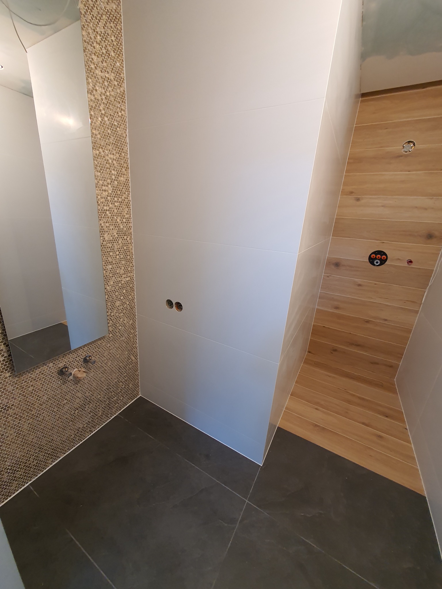 Nowoczesna łazienka w trakcie wykończenia: szare płytki podłogowe, ściany z białych, gładkich płytek, fragment ściany z mozaiki przy lustrze, drewnopodobne płytki na ścianie z instalacją hydrauliczną.