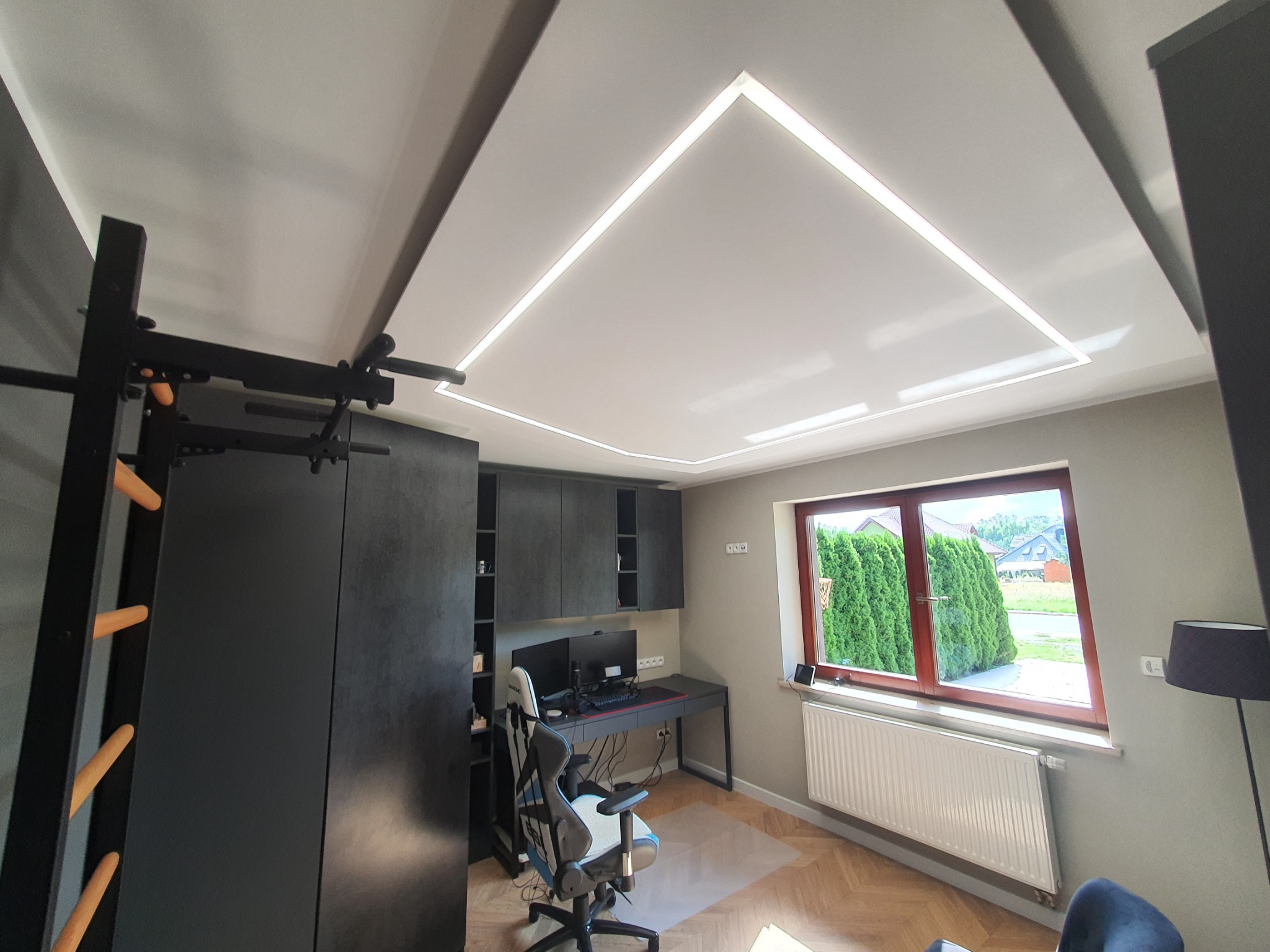 Pokój z nowoczesnym oświetleniem sufitowym w kształcie kwadratu, biurkiem z dwoma monitorami, drabinką gimnastyczną i parkietem ułożonym we wzór jodełki francuskiej.