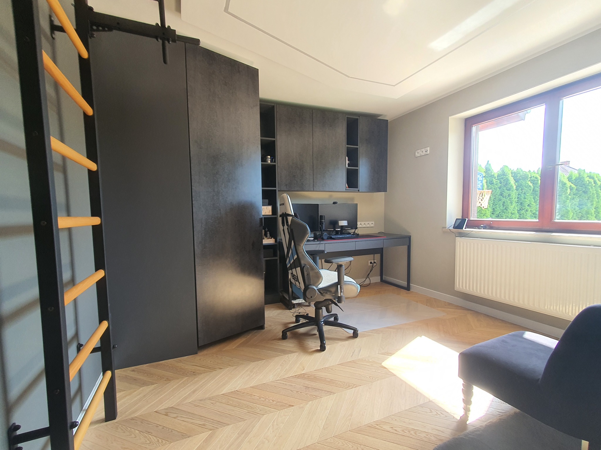 Pokój z parkietem w jodełkę, zabudową meblową w kolorze grafitowym, drabinką gimnastyczną, biurkiem i krzesłem gamingowym, oświetlony promieniami słonecznymi wpadającymi przez okno.