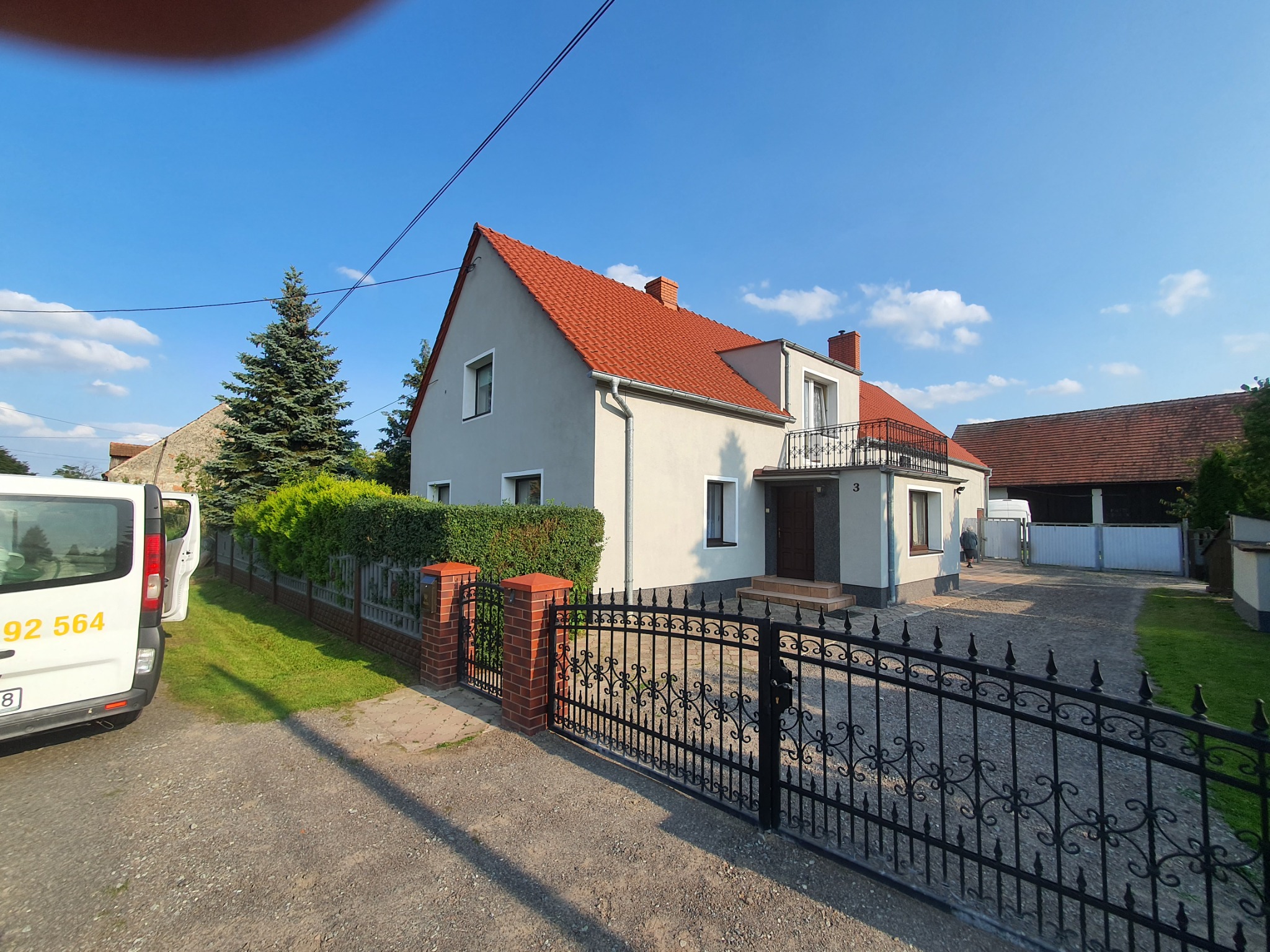 Dwupiętrowy dom z czerwonym dachem i balkonem, widoczny zza ozdobnego, czarnego ogrodzenia, z zaparkowanym białym vanem z numerem telefonu po lewej stronie.