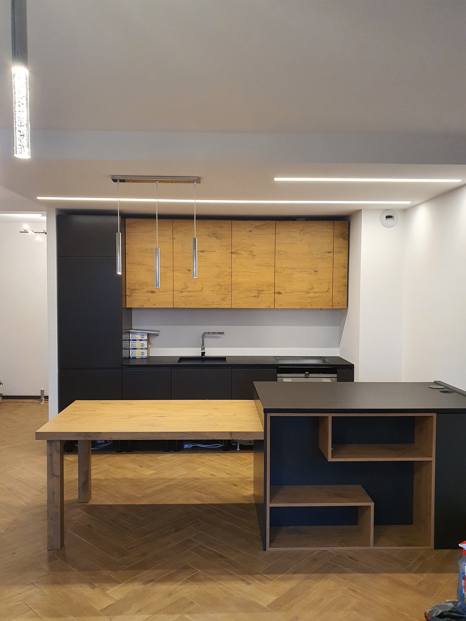 Nowoczesna kuchnia z drewnianymi akcentami: czarne szafki dolne i wyspa, górne szafki z forniru imitującego drewno, parkiet w jodełkę, minimalistyczne oświetlenie LED.