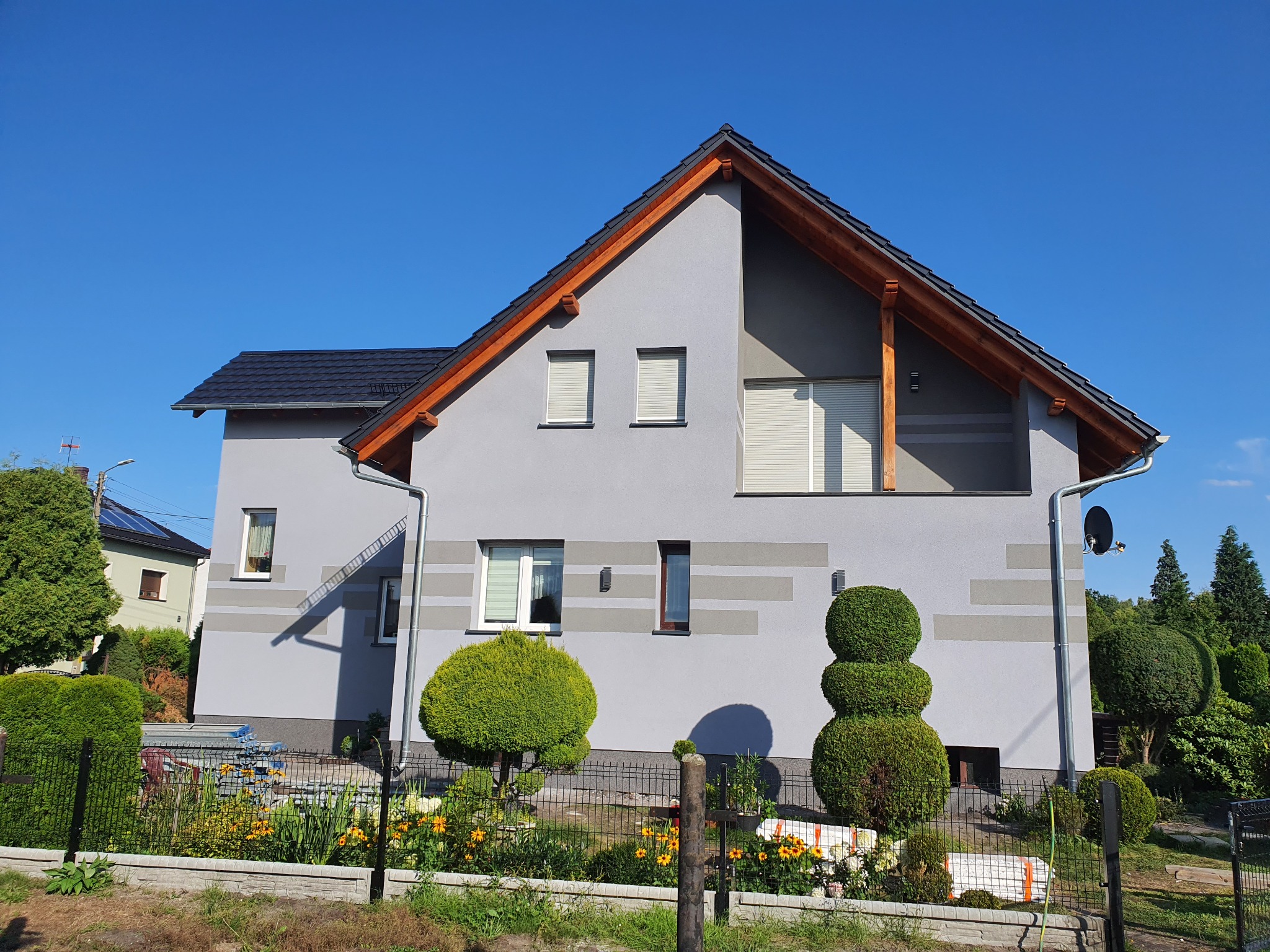 Szary dom jednorodzinny z ciemną dachówką i drewnianymi elementami wykończeniowymi, widok z ogrodu z ozdobnymi krzewami