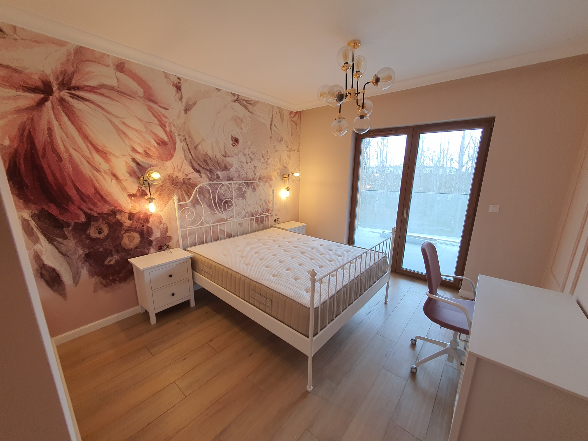 Sypialnia z łóżkiem na białej, metalowej ramie, kwiecista tapeta z motywem róż, stoliki nocne z lampkami, nowoczesna, okrągła lampa wisząca i biurko z krzesłem.