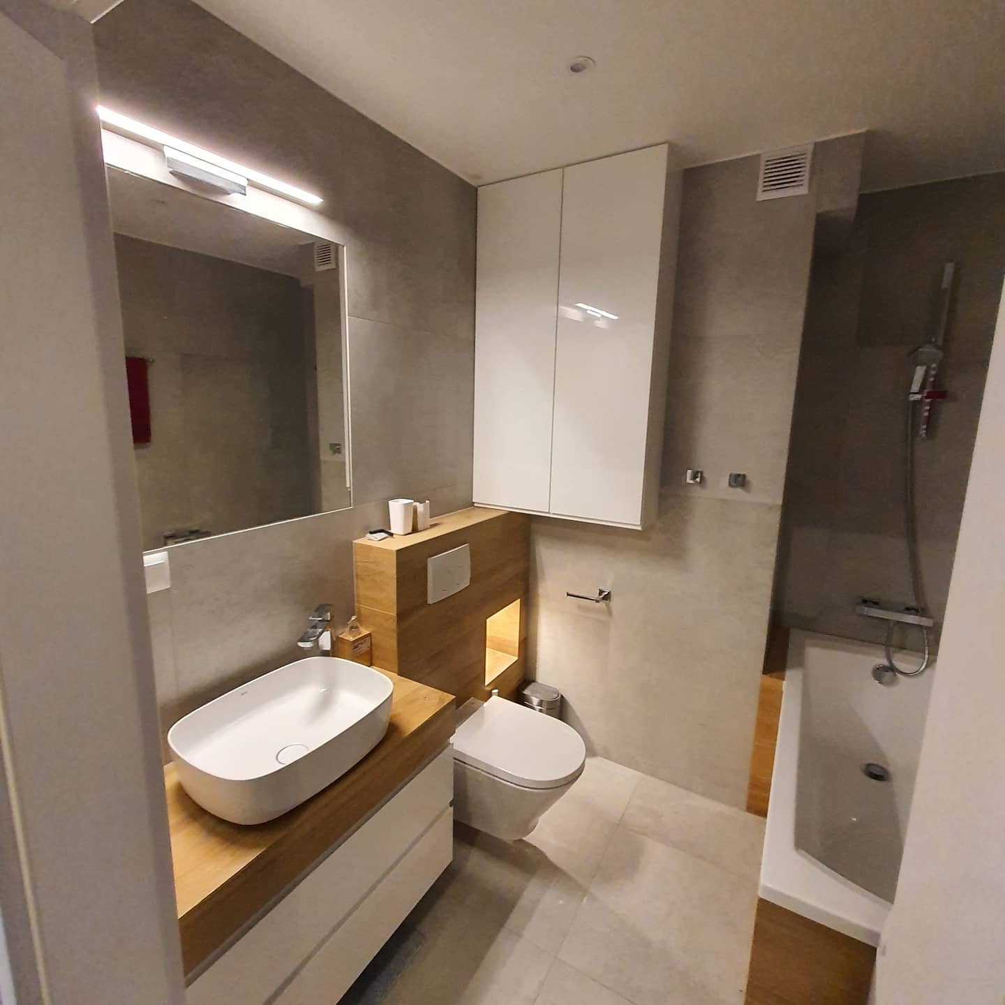 Nowoczesna łazienka w szarościach z drewnianymi akcentami: umywalka nablatowa, zabudowa toalety z podświetleniem, białe szafki wiszące i wanna z prysznicem.