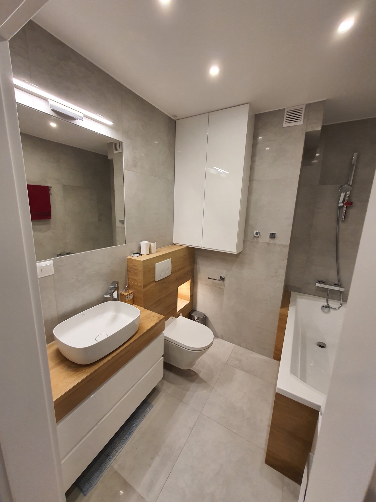 Nowoczesna łazienka w odcieniach szarości i bieli z drewnianymi akcentami, owalna umywalka nablatowa, podwieszana toaleta i wanna z prysznicem.