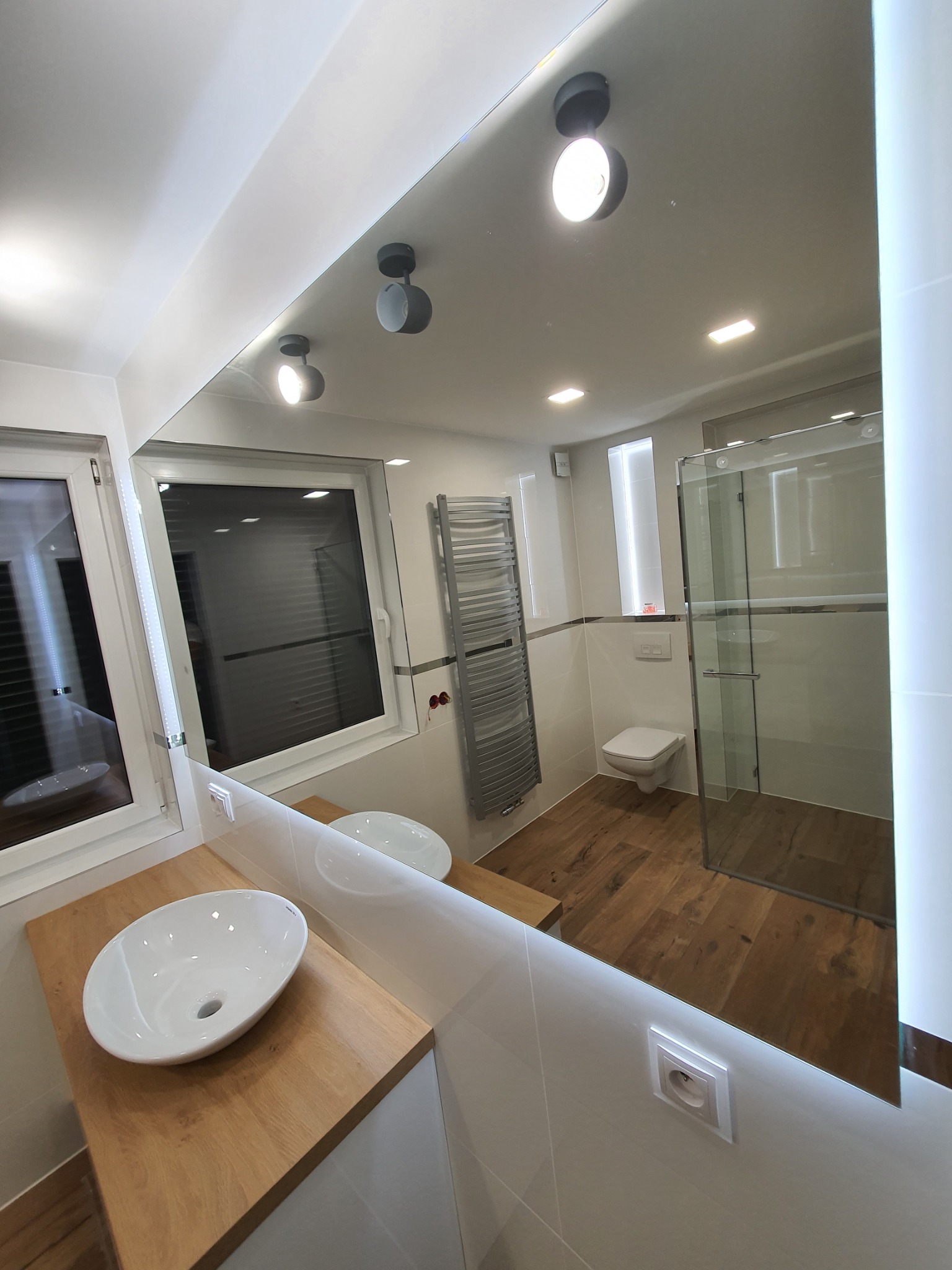 Nowoczesna łazienka z drewnianą podłogą, kabiną prysznicową ze szkła i umywalką nablatową, odbicie w lustrze z podświetleniem LED.