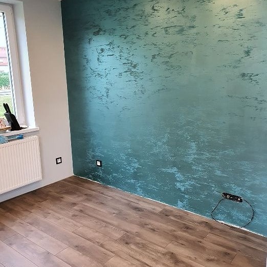 Turkusowa ściana z efektem dekoracyjnym, drewnopodobne panele podłogowe, biały grzejnik pod oknem, widoczne gniazdka elektryczne.