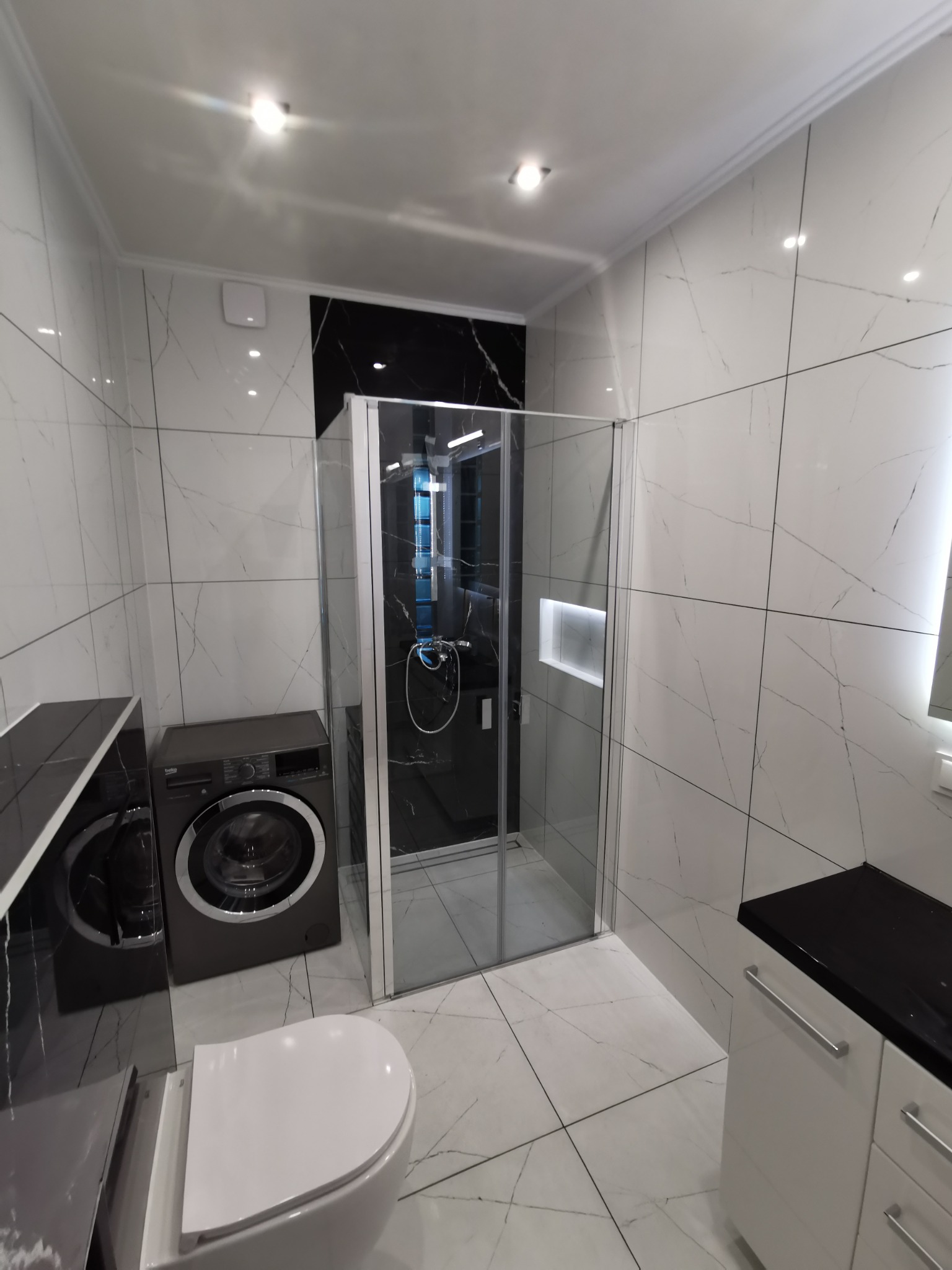 Nowoczesna łazienka z białymi płytkami imitującymi marmur, kabina prysznicowa z czarnym panelem, pralka i szafka z czarnym blatem.