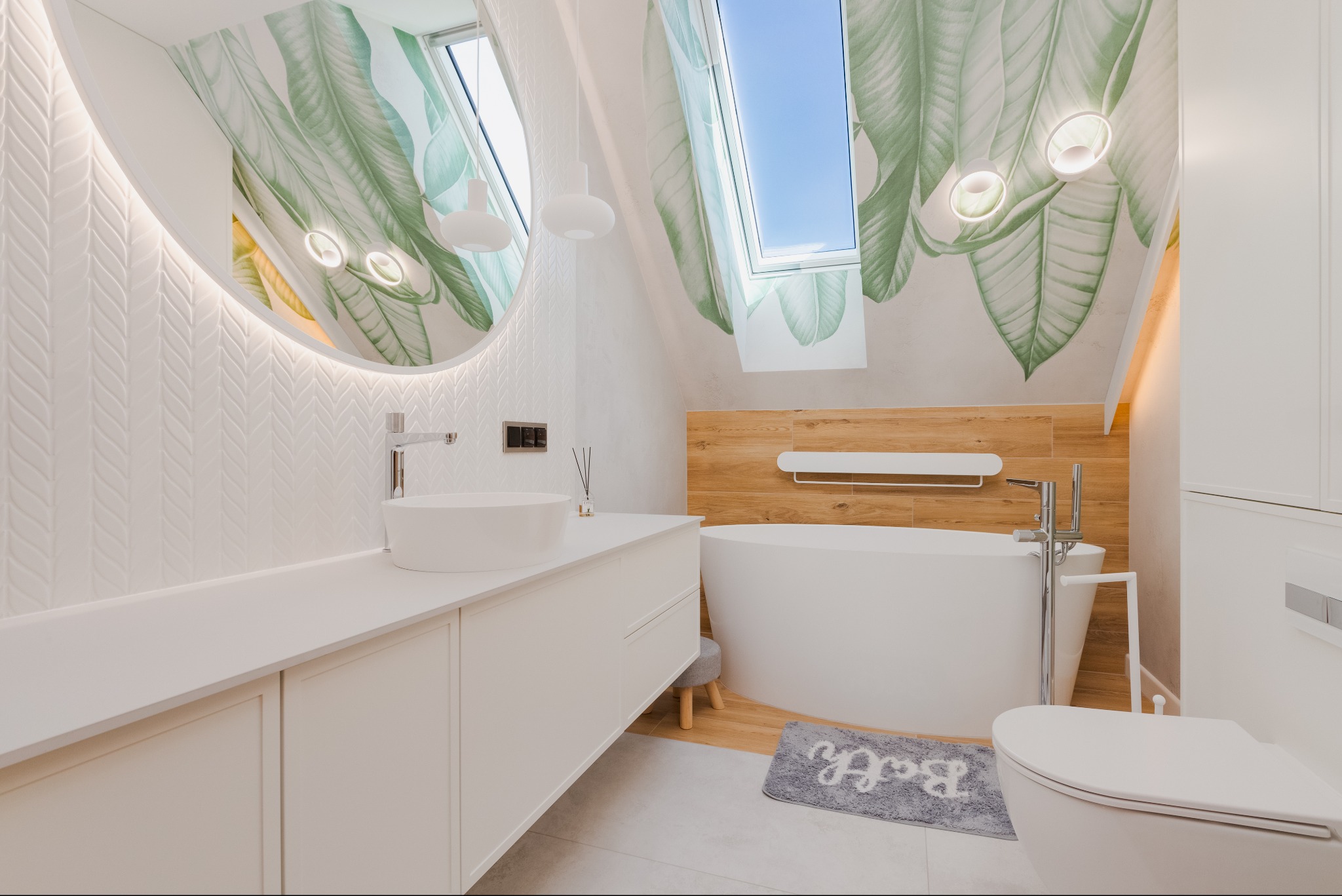 Jasna łazienka na poddaszu z owalnym lustrem z podświetleniem LED, białą umywalką nablatową i wanną wolnostojącą na tle ściany z drewnopodobną okładziną oraz tapetą w motywy roślinne.