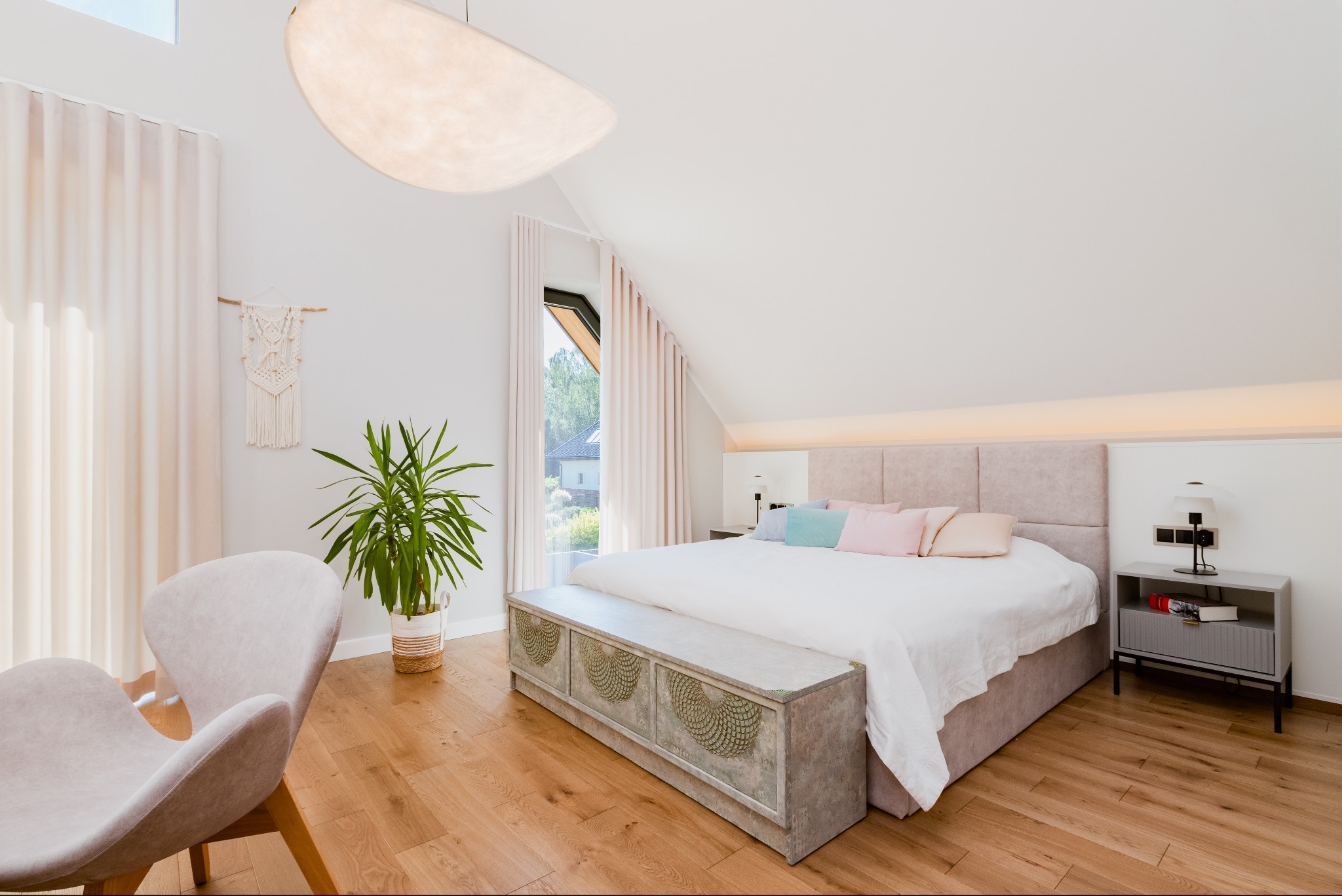 Jasna sypialnia na poddaszu z drewnianą podłogą, beżowym łóżkiem z pikowanym zagłówkiem, ozdobną skrzynią u podnóża, designerską lampą sufitową i fotelem w rogu.
