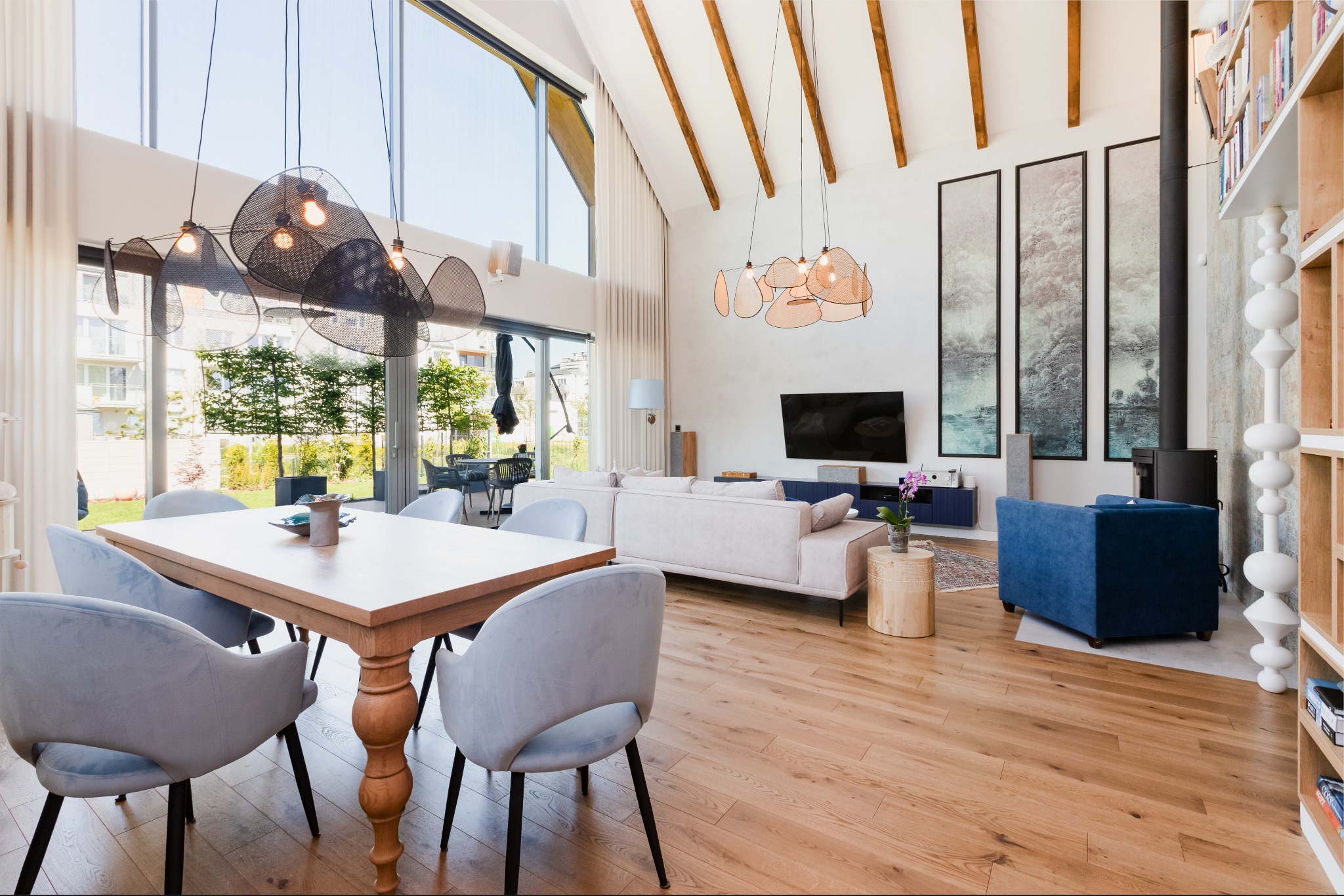 Jasne, przestronne wnętrze domu z drewnianą podłogą, stołem jadalnym z szarymi krzesłami, sofą, błękitnym fotelem i dużymi oknami z widokiem na ogród.