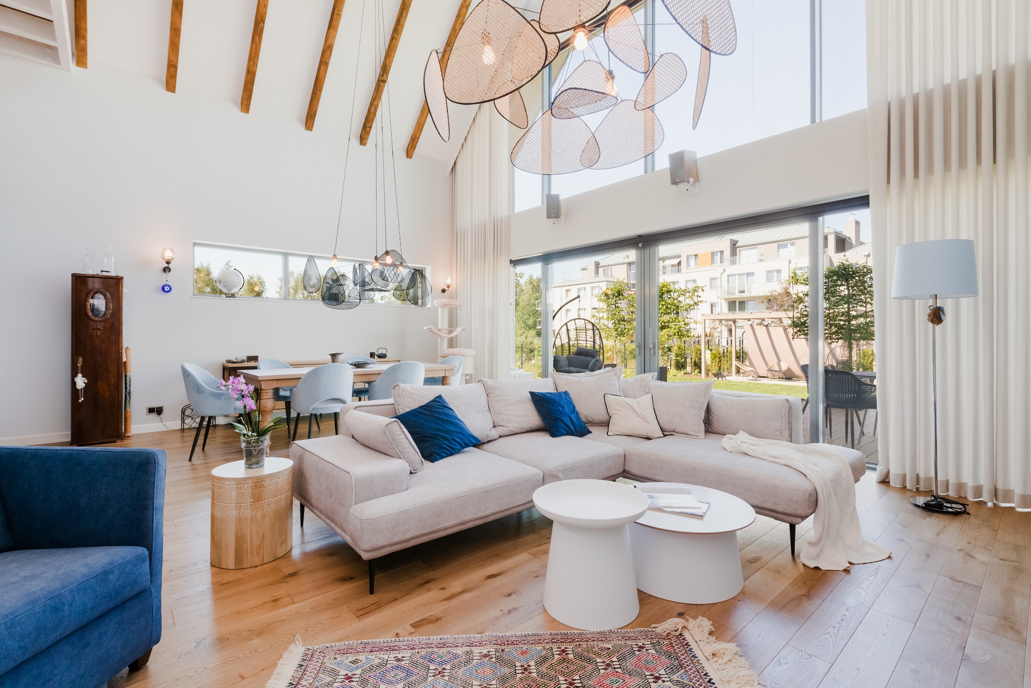 Jasne, przestronne wnętrze salonu z wysokim sufitem, drewnianą podłogą, narożną sofą w odcieniach beżu i niebieskimi poduszkami, oraz designerską lampą wiszącą i dużymi oknami z widokiem na ogród.